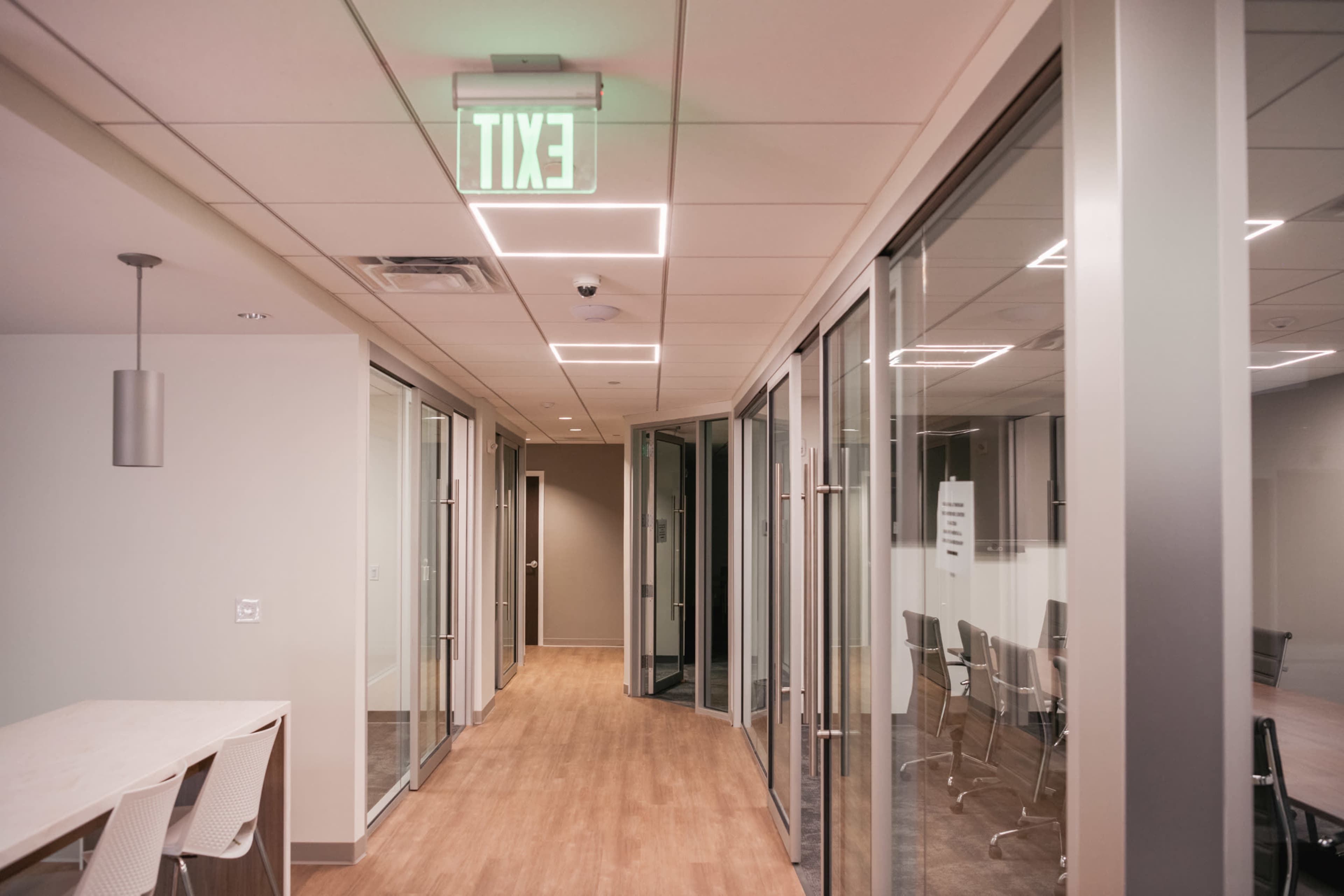 The image shows a modern office corridor lined with glass-walled meeting rooms and an illuminated exit sign.