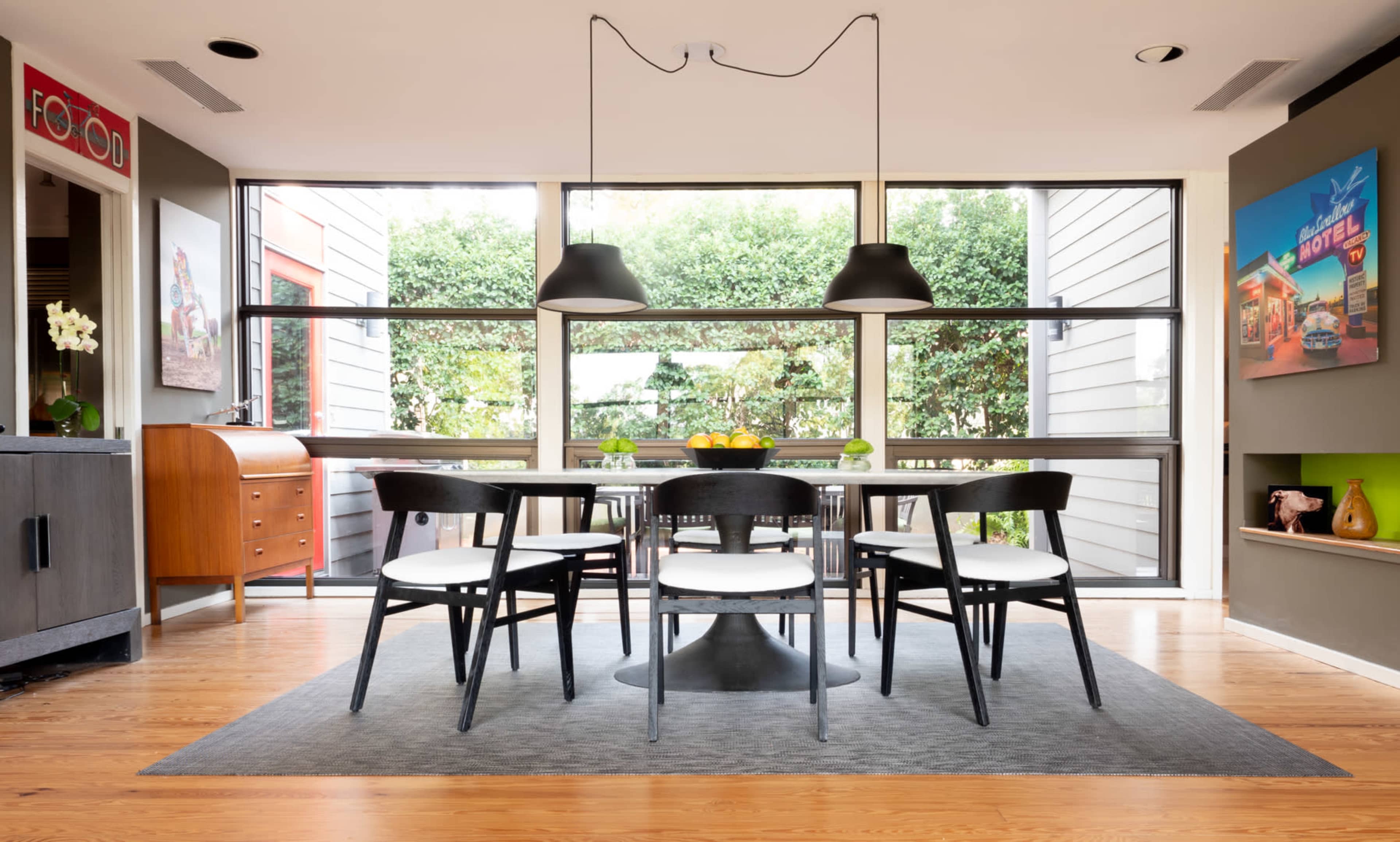 A modern dining area features a round table surrounded by six black chairs, with large windows allowing natural light and a view of greenery.