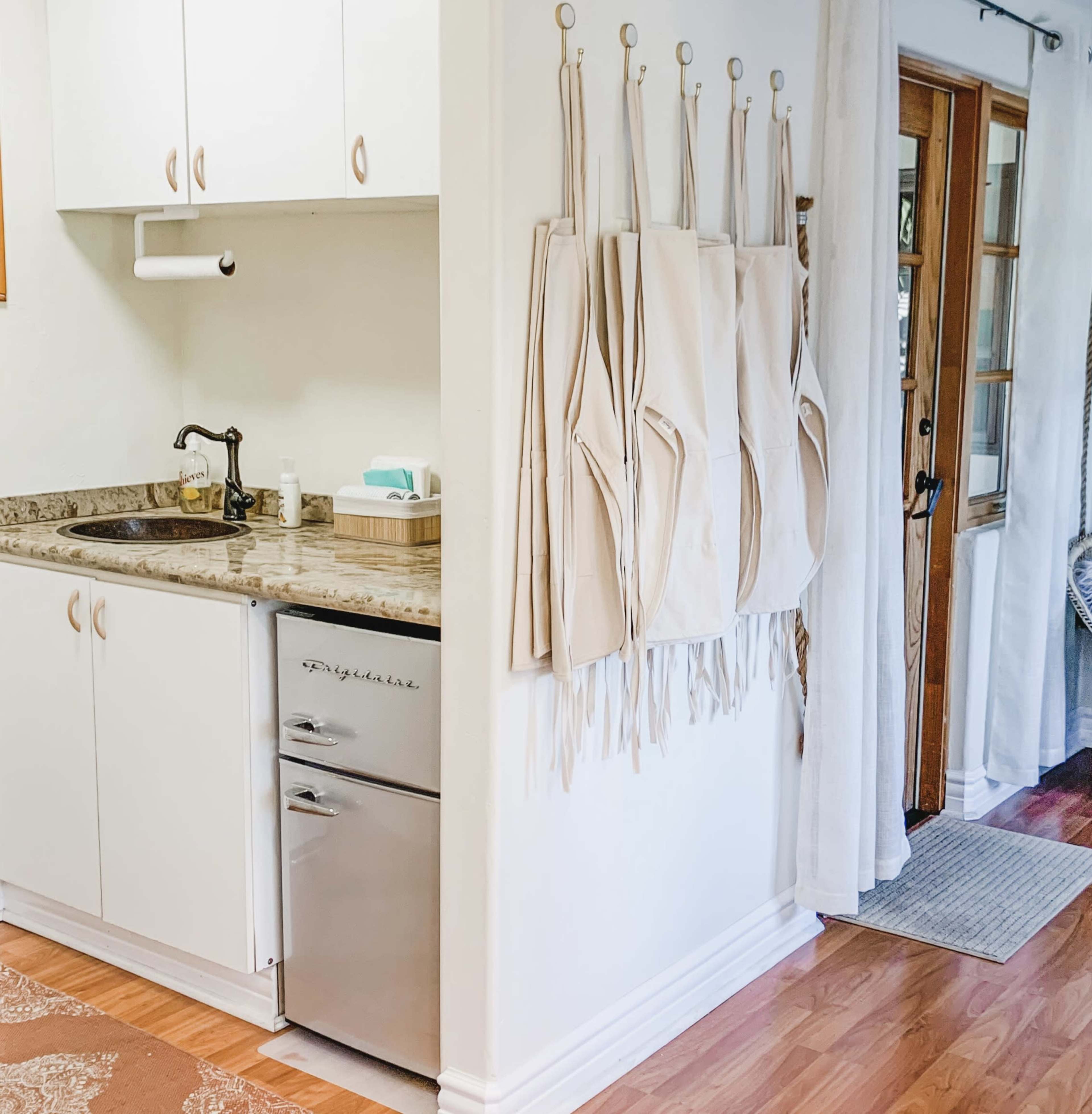 A kitchen area with a sink, a small refrigerator, and several beige aprons hanging on the wall.