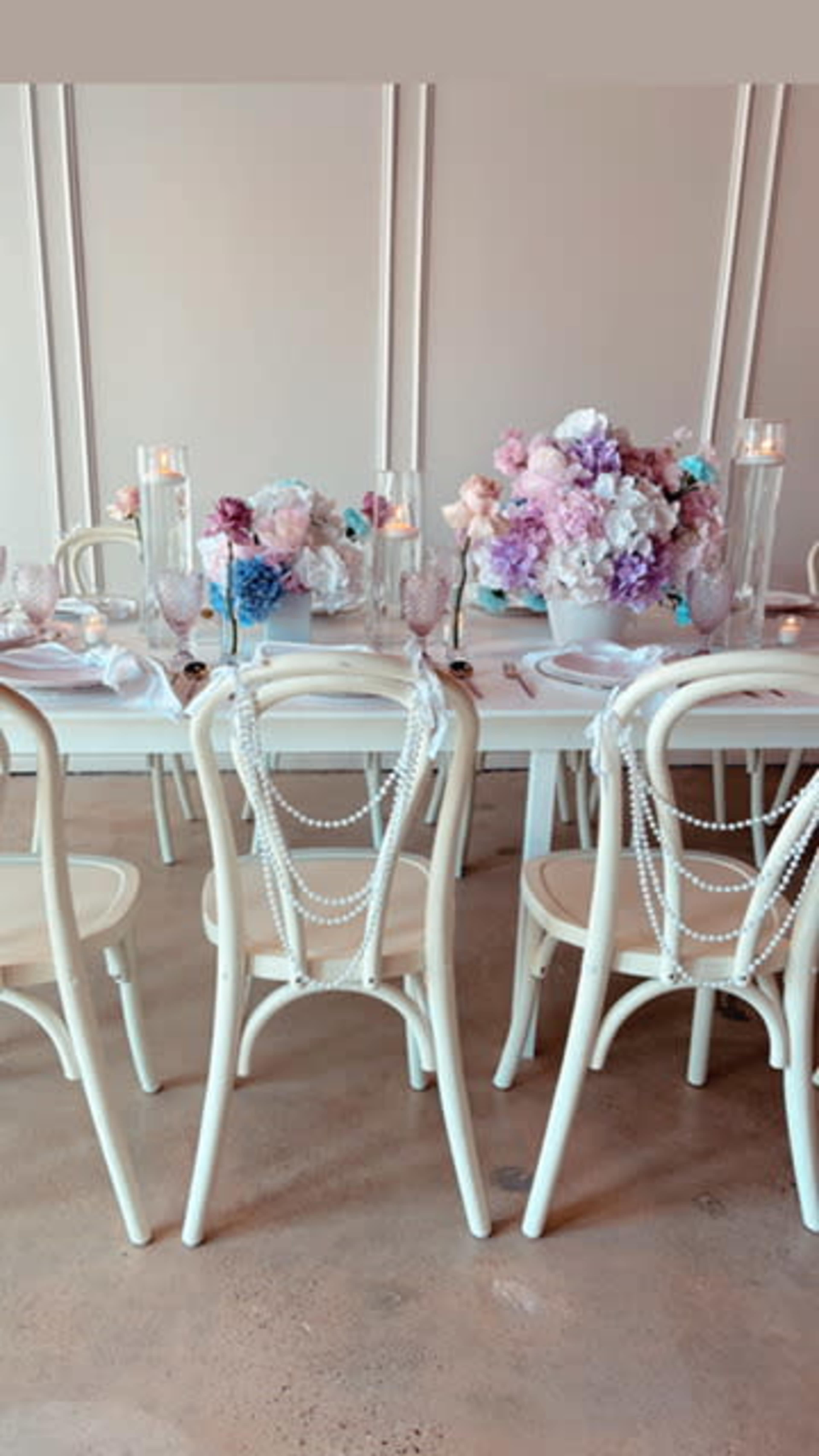 A beautifully set dining table features white chairs adorned with pearl strands and a centerpiece of pink and purple flowers.