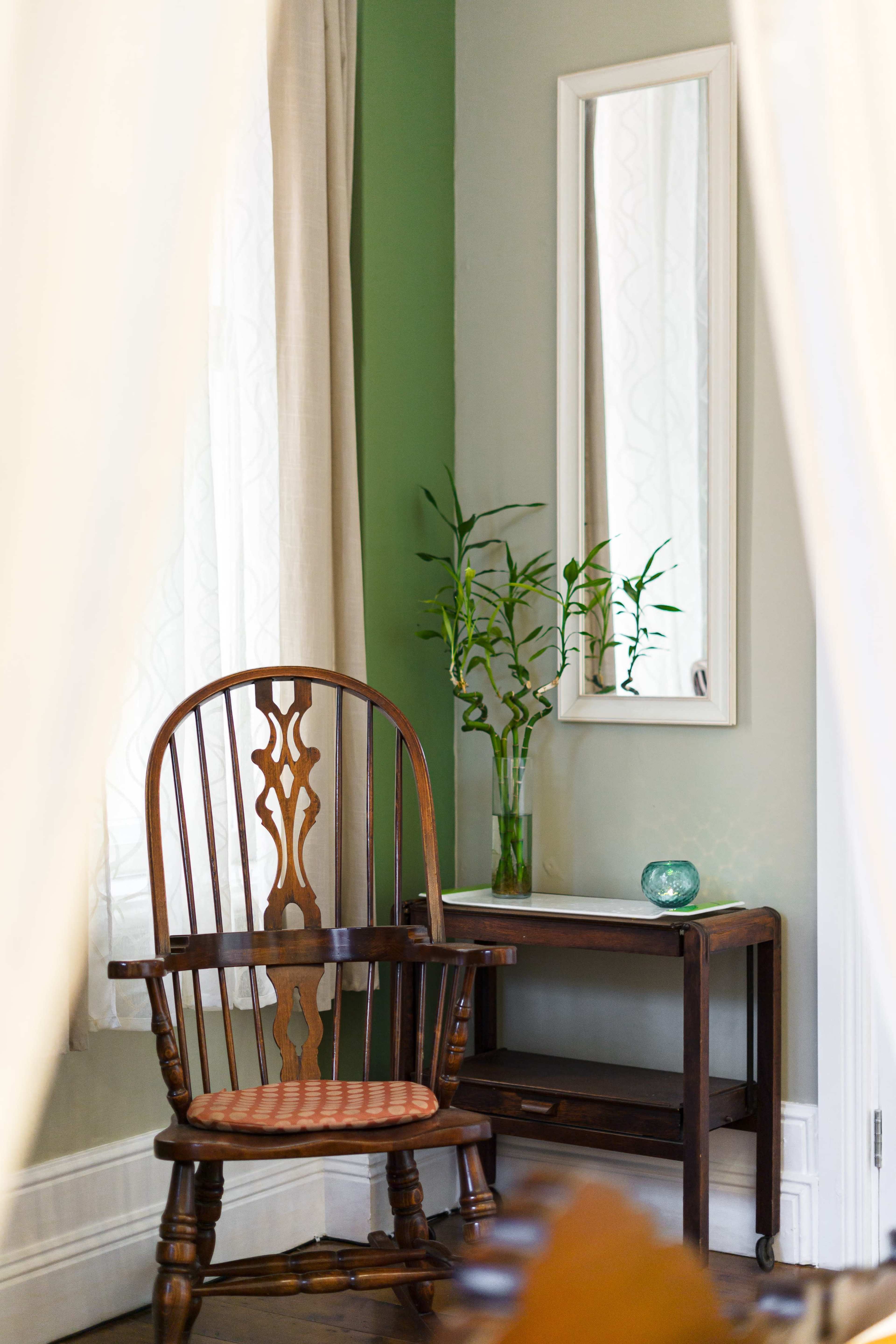 A wooden rocking chair is positioned near a table with a small vase of bamboo and a mirror on the wall.