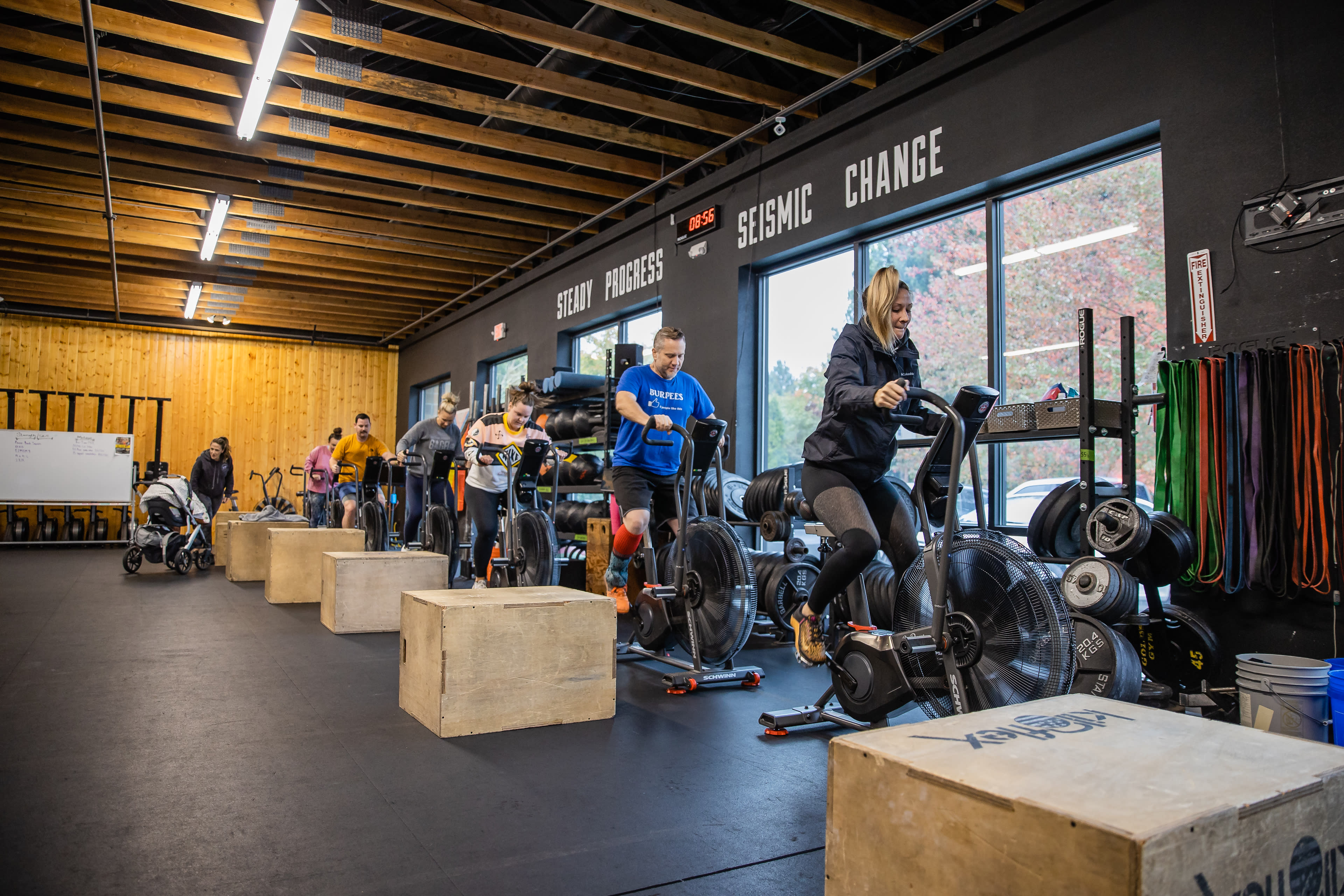 A group of fitness participants are exercising on stationary bikes in a gym with wooden flooring and various workout equipment.