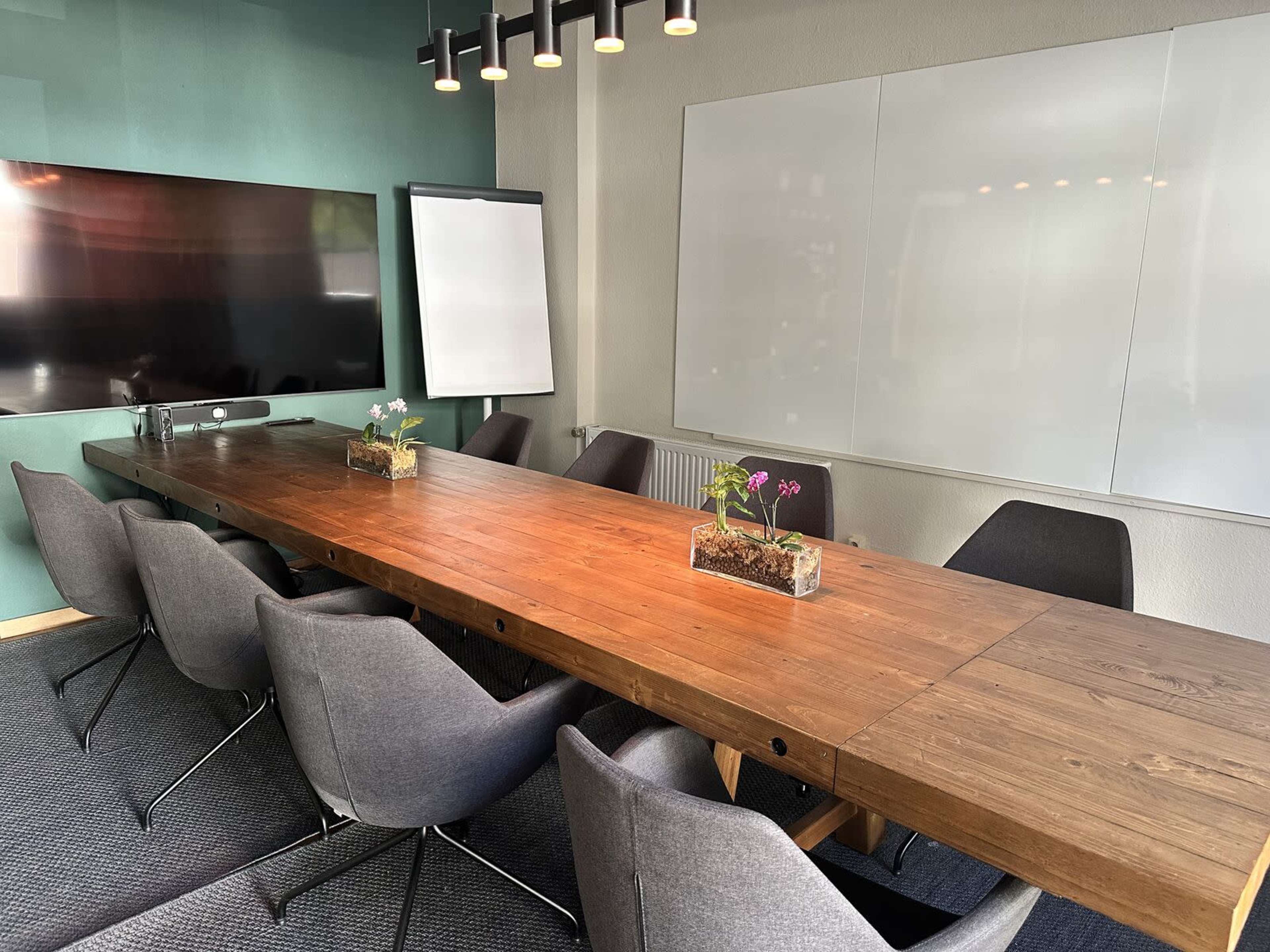 The image shows a modern conference room featuring a long wooden table surrounded by gray chairs, a flat-screen TV, a whiteboard, and potted plants on the table.