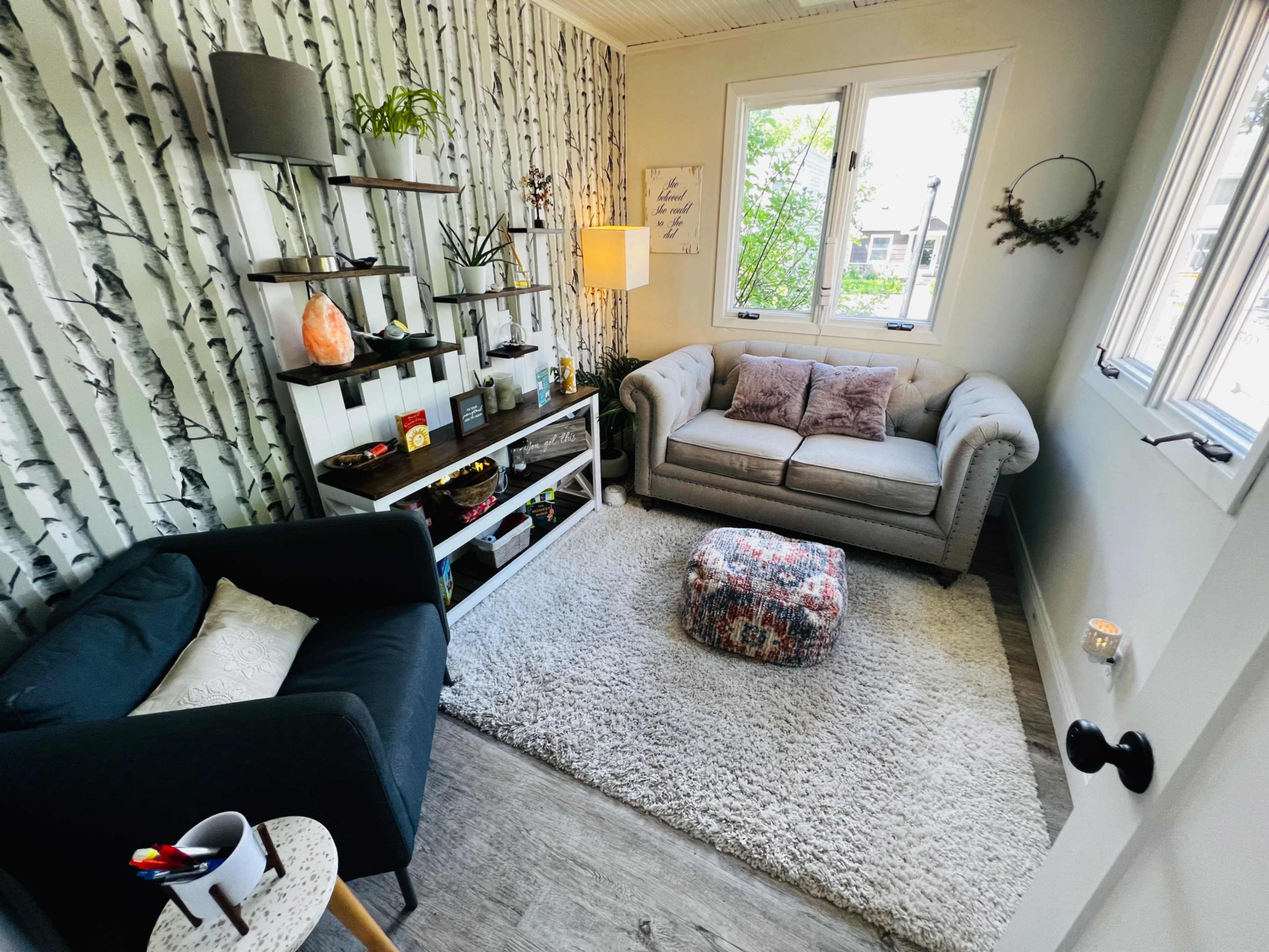 A cozy room featuring a gray sofa, a black chair, a patterned ottoman, and shelves with decorative items against a birch tree-patterned wall.