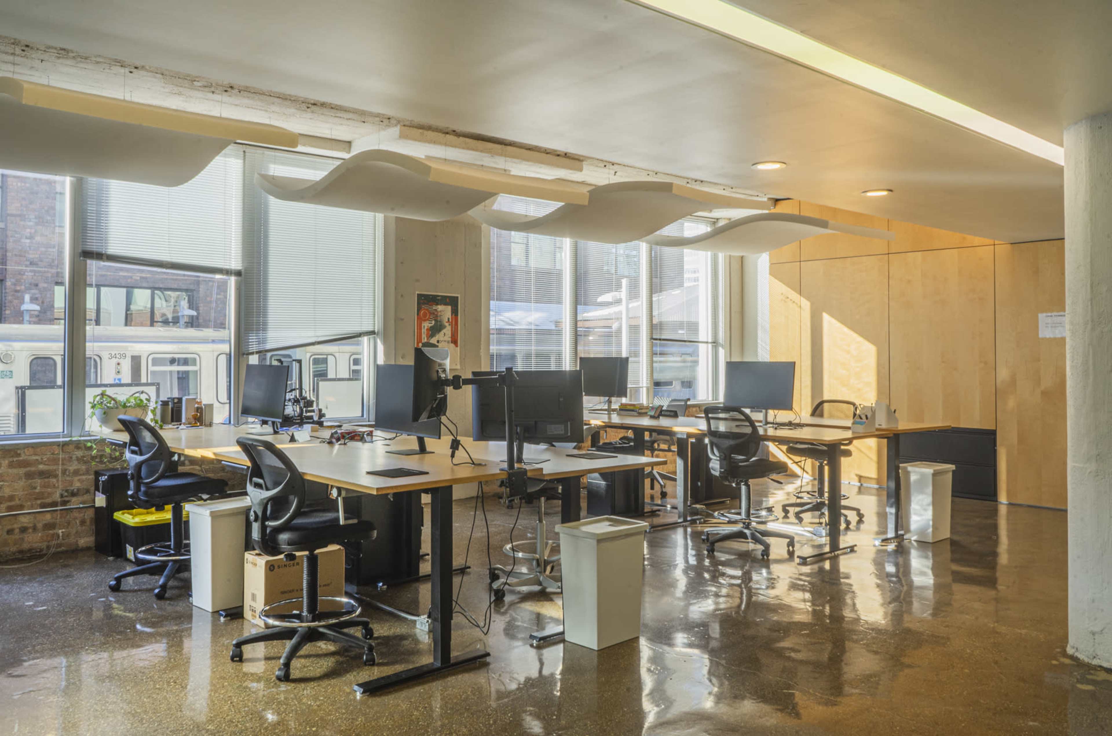 A bright office space features multiple workstations with desks, computers, and ergonomic chairs, illuminated by large windows.
