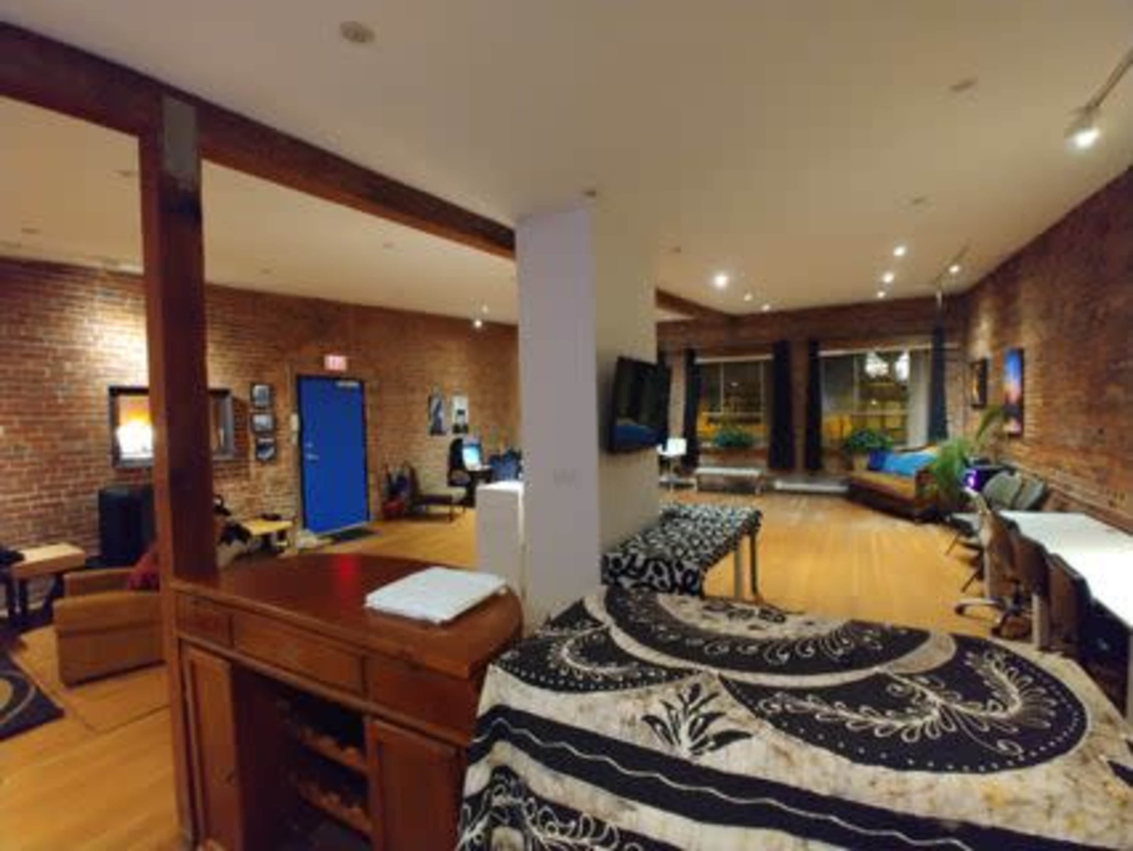 The image shows a spacious room with exposed brick walls, wooden flooring, and various furniture pieces, including a bed, couches, and workstations.