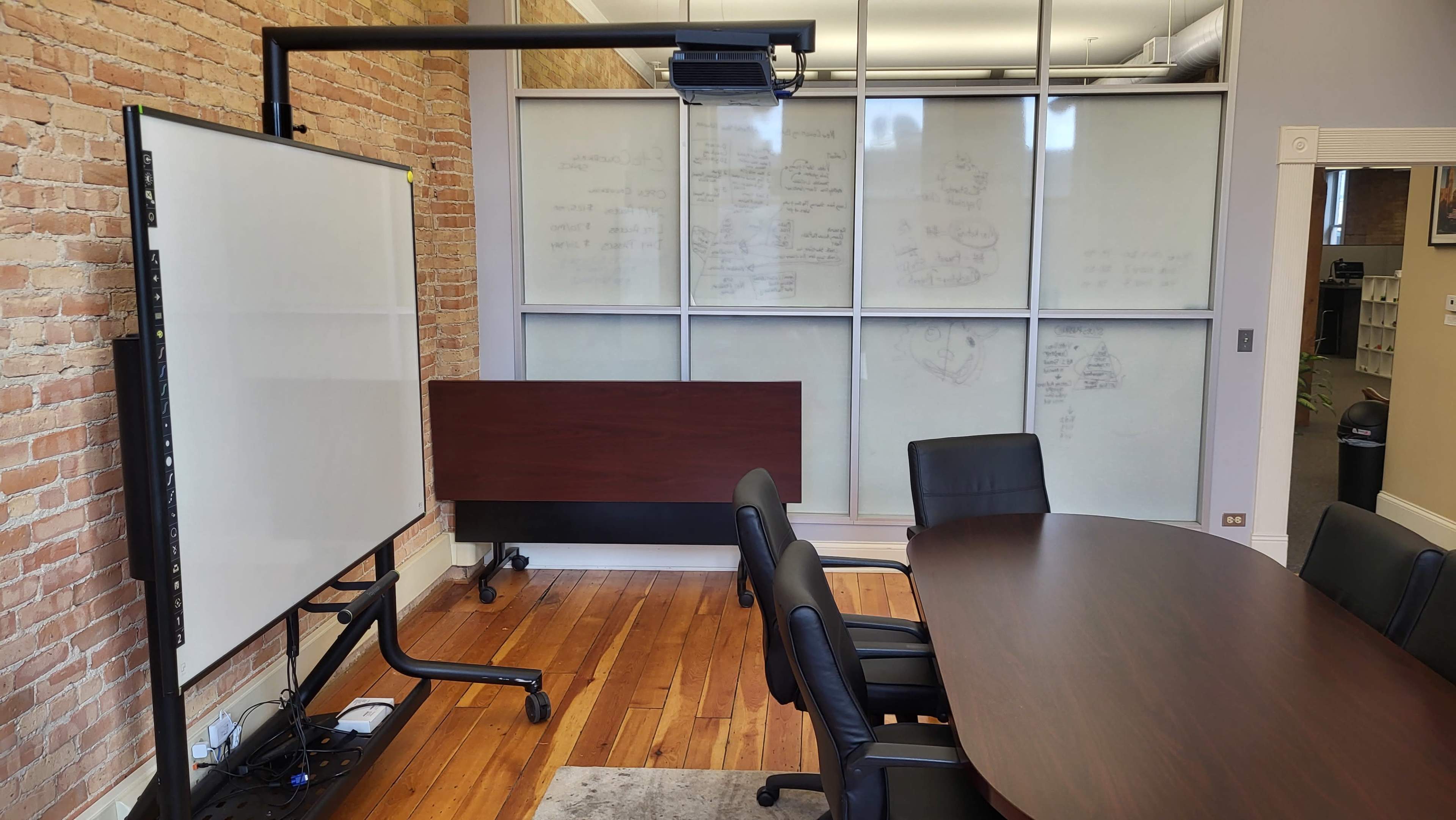 A conference room features a large oval table surrounded by black chairs, with a whiteboard on one side and a glass wall displaying whiteboard notes.
