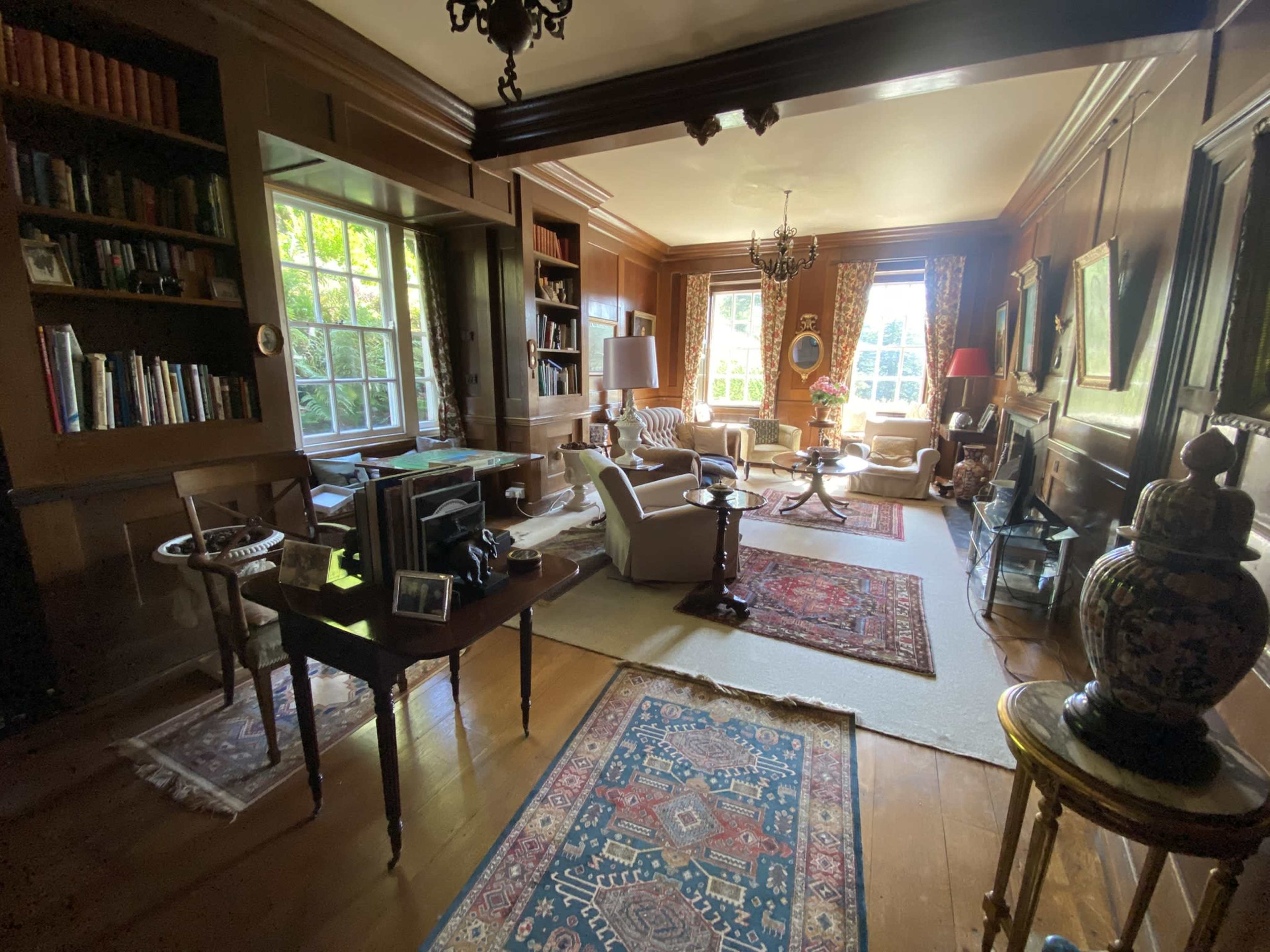 The image shows a cozy living room with wooden paneling, bookshelves, several armchairs, a coffee table, and large windows letting in natural light.