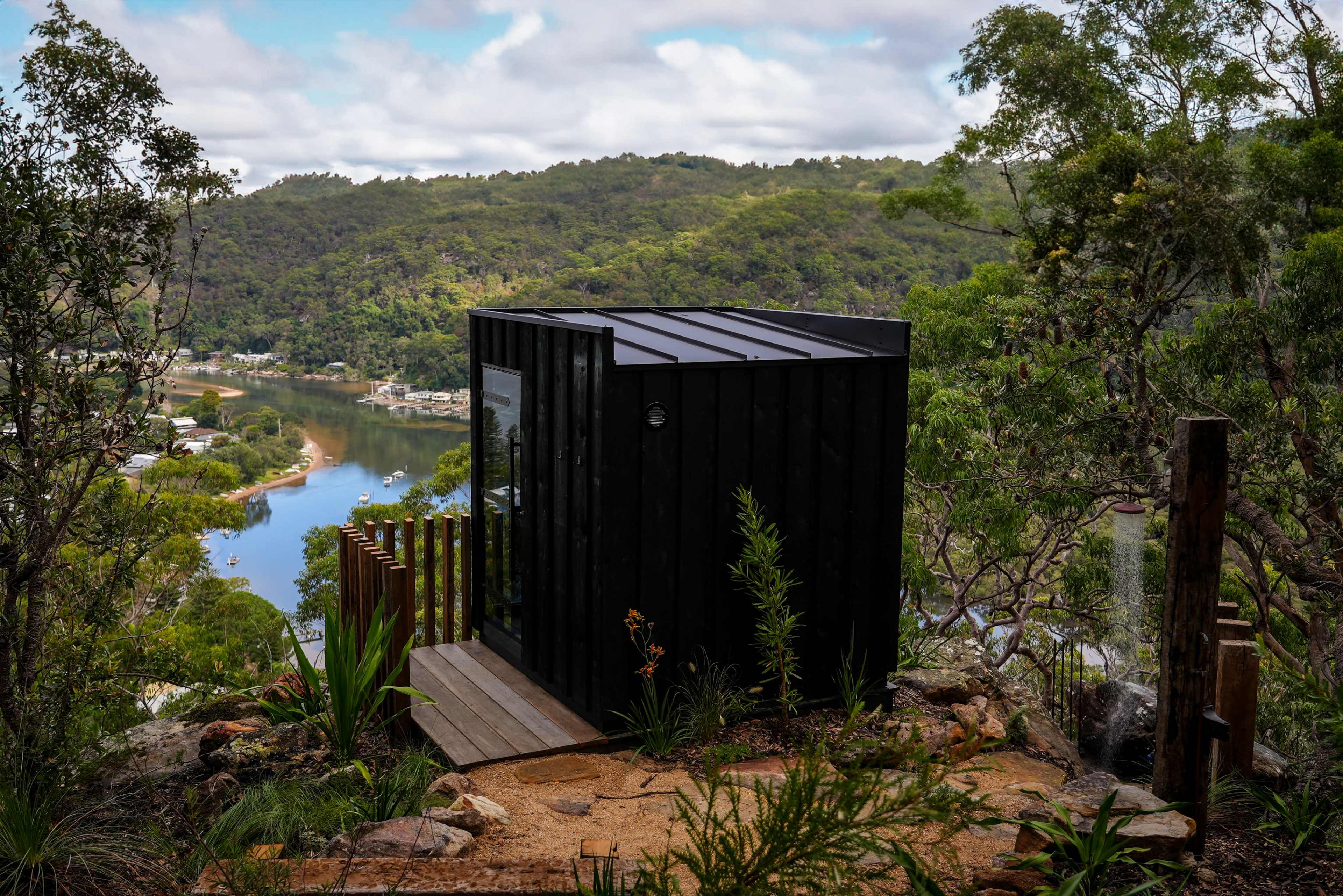 A small black cabin is situated on a hillside overlooking a river and lush green mountains.