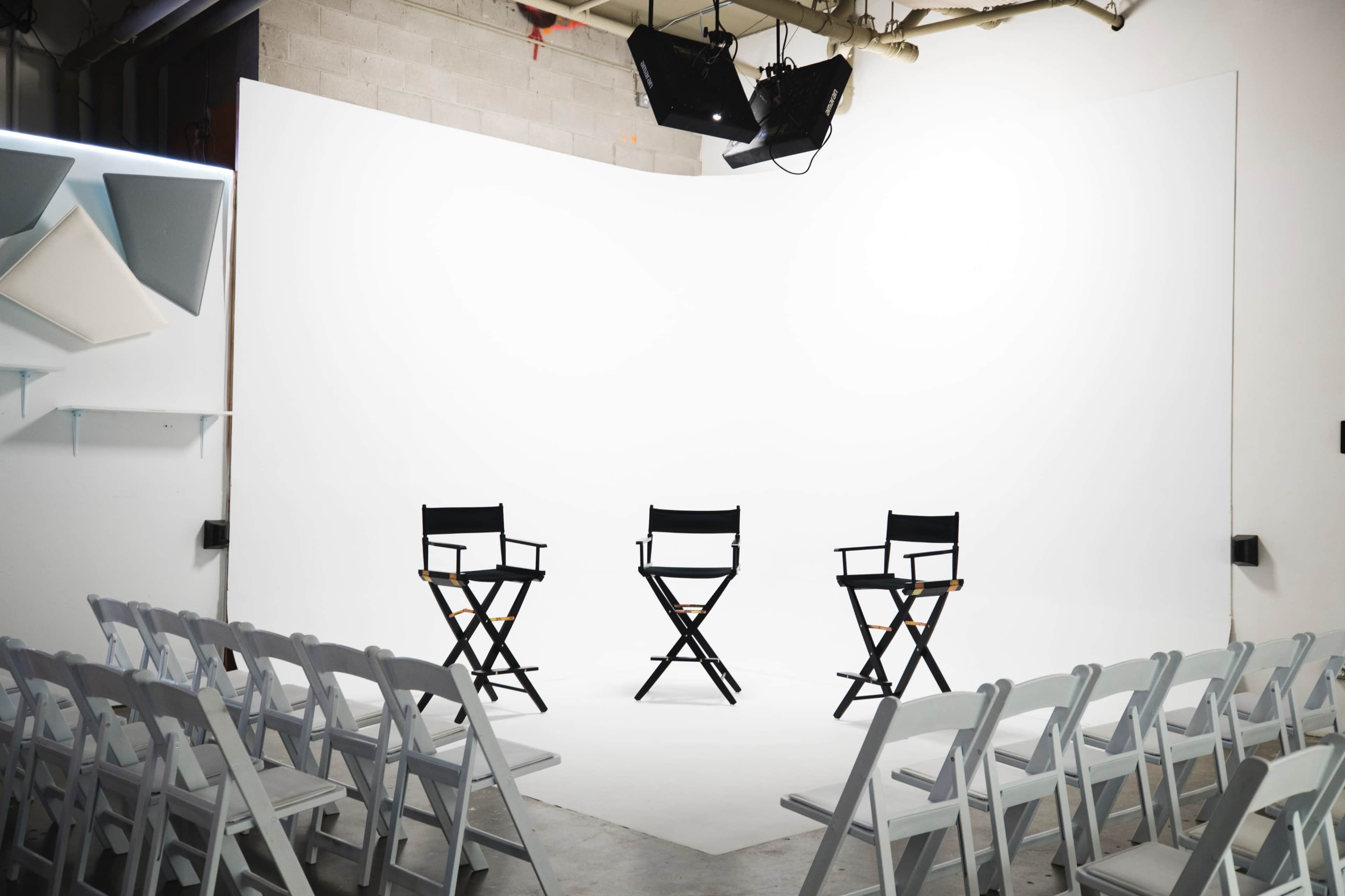 The image shows a studio setup with three director's chairs facing an empty audience of white folding chairs in front of a plain white backdrop.