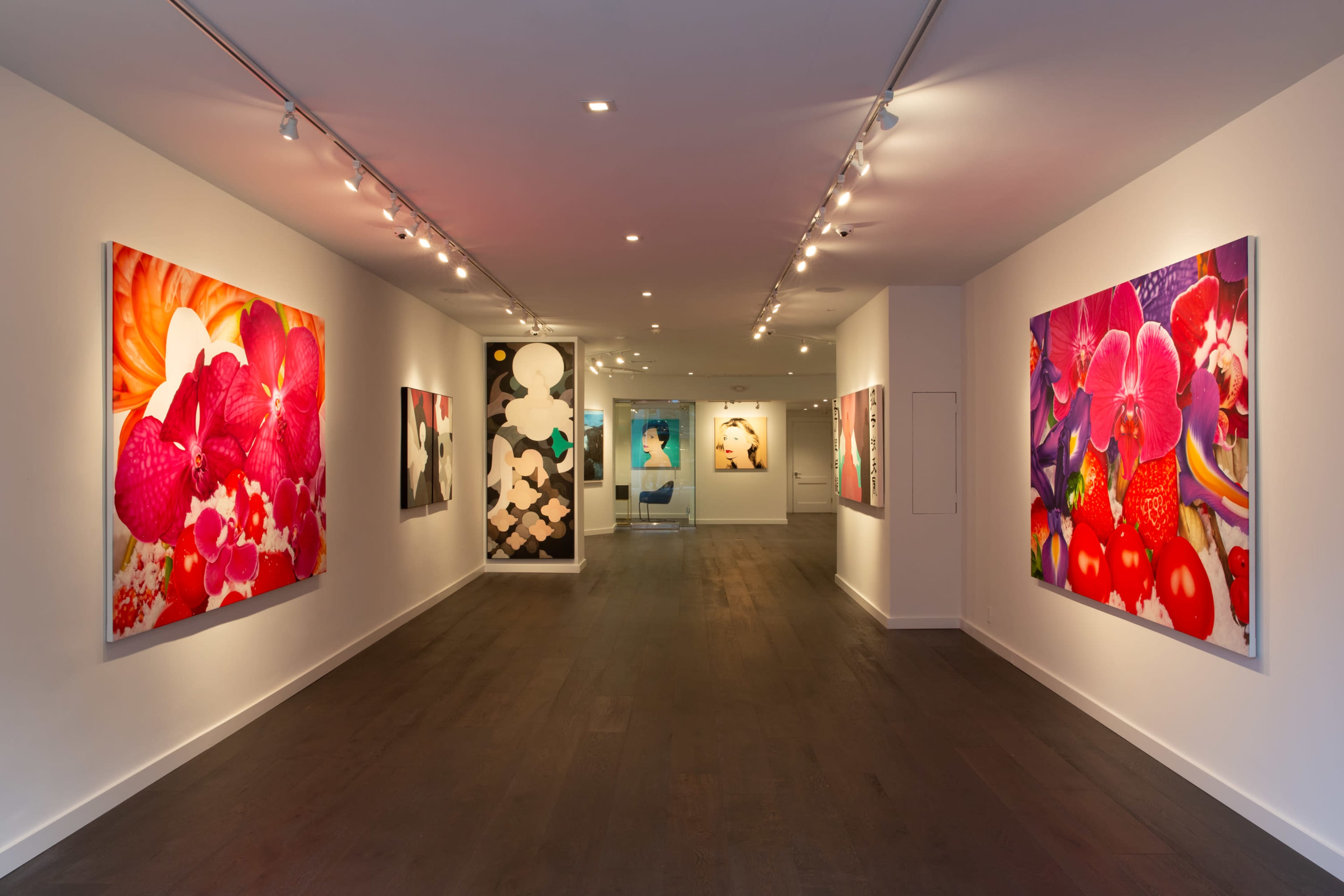 A brightly lit art gallery features large, colorful paintings of flowers and abstract designs on the walls.