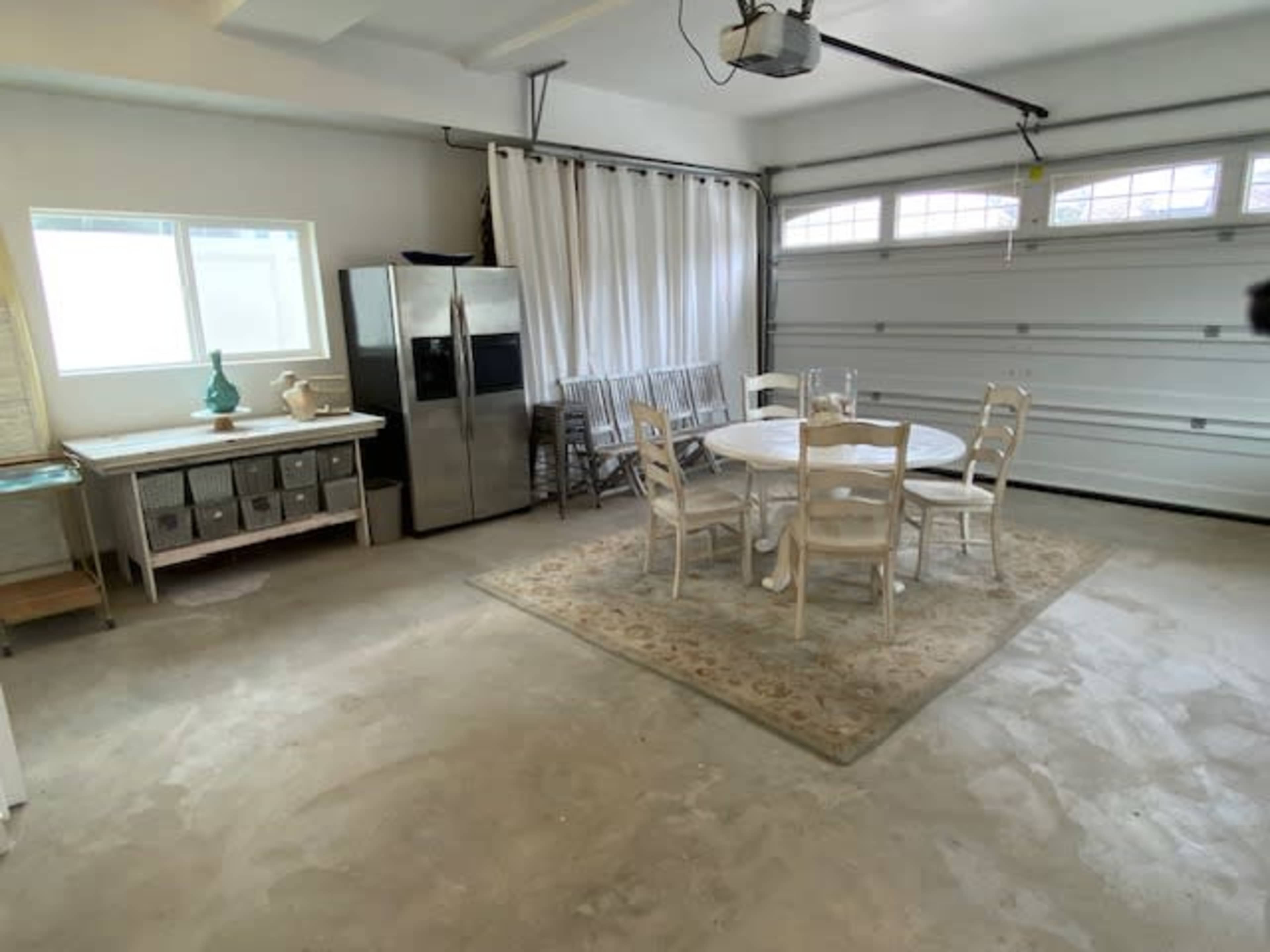 The image shows a garage that has been converted into a dining area, featuring a round table with chairs, a rug, and a refrigerator along with storage bins.
