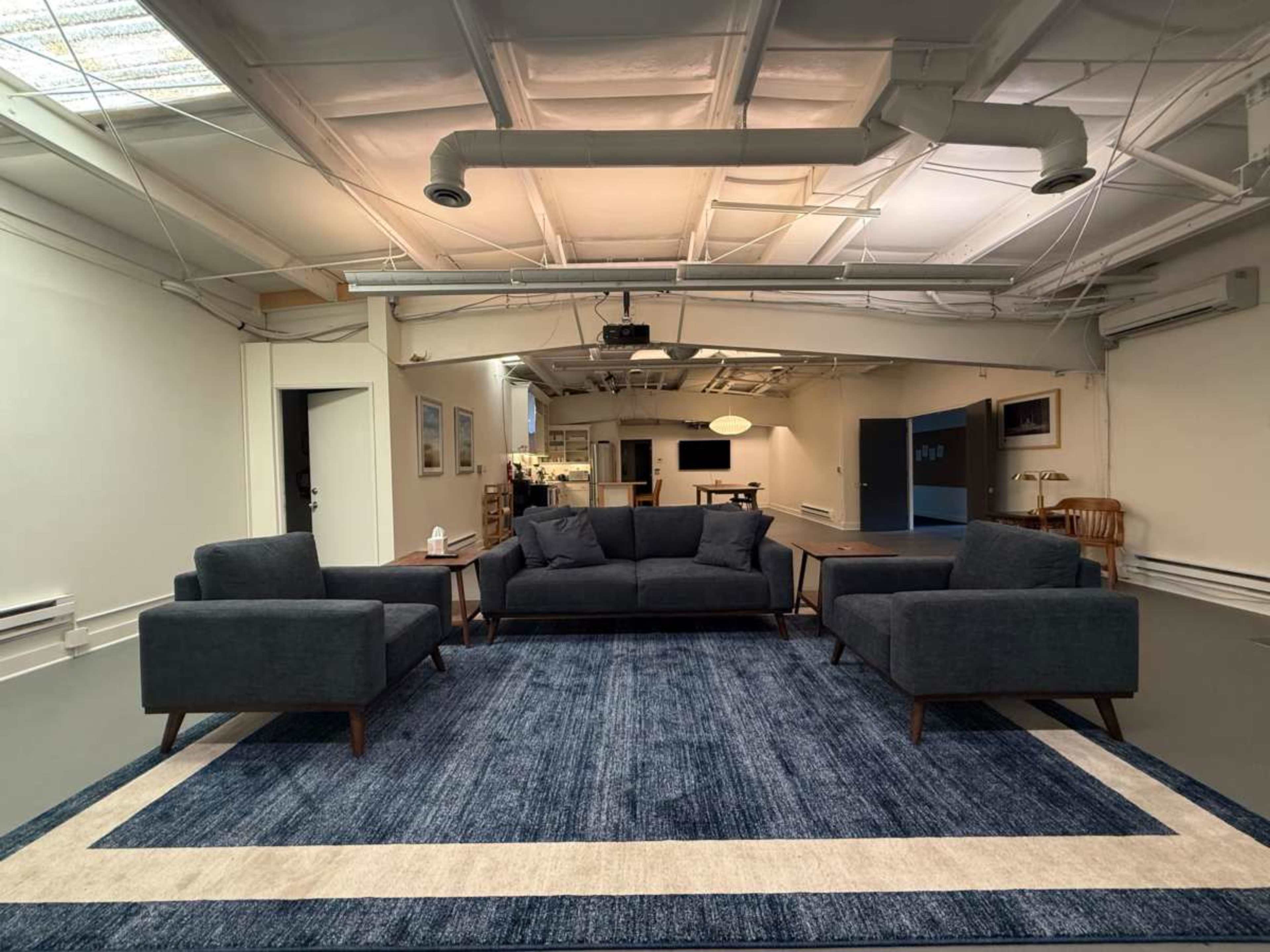 The image shows a spacious room with a large, dark blue area rug centered beneath a sectional sofa and two armchairs, all arranged in a minimalist style.