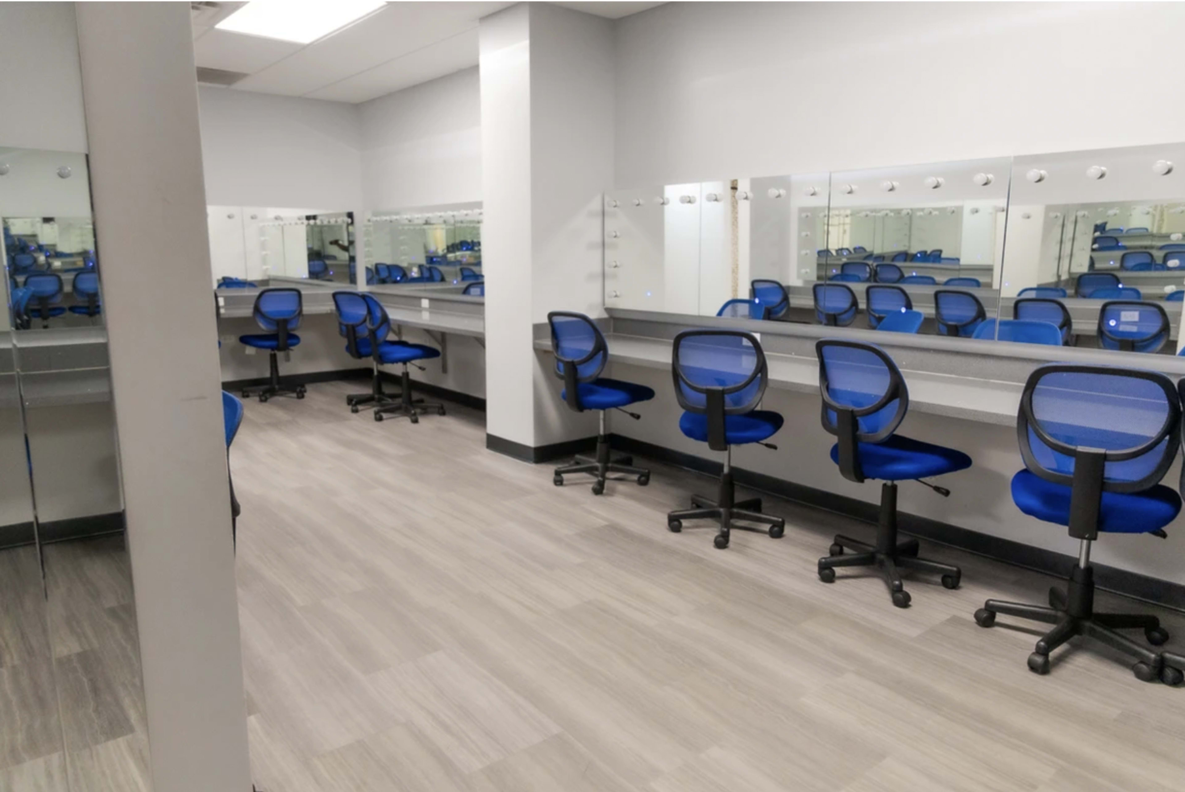 A brightly lit dressing room with several grey countertops and blue rolling chairs arranged in front of mirrors.