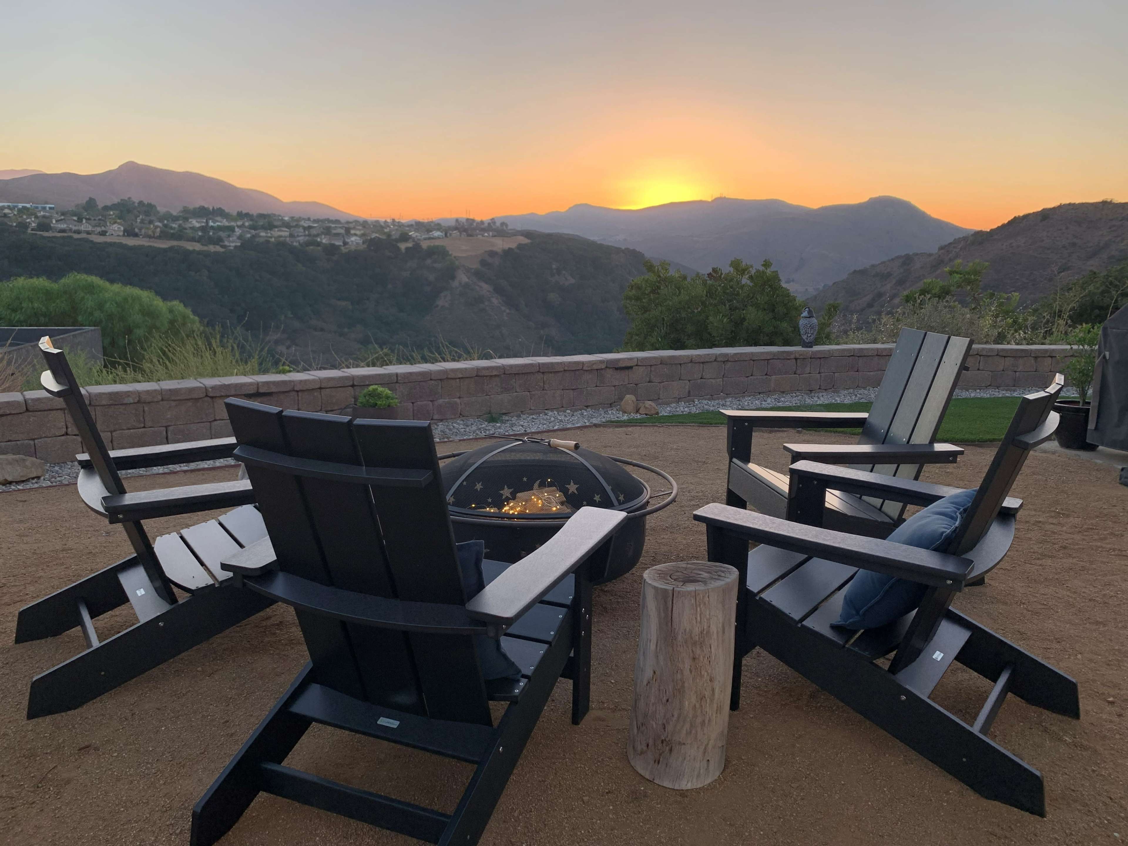 Four black Adirondack chairs are arranged around a fire pit, set against a backdrop of mountains and a sunset.
