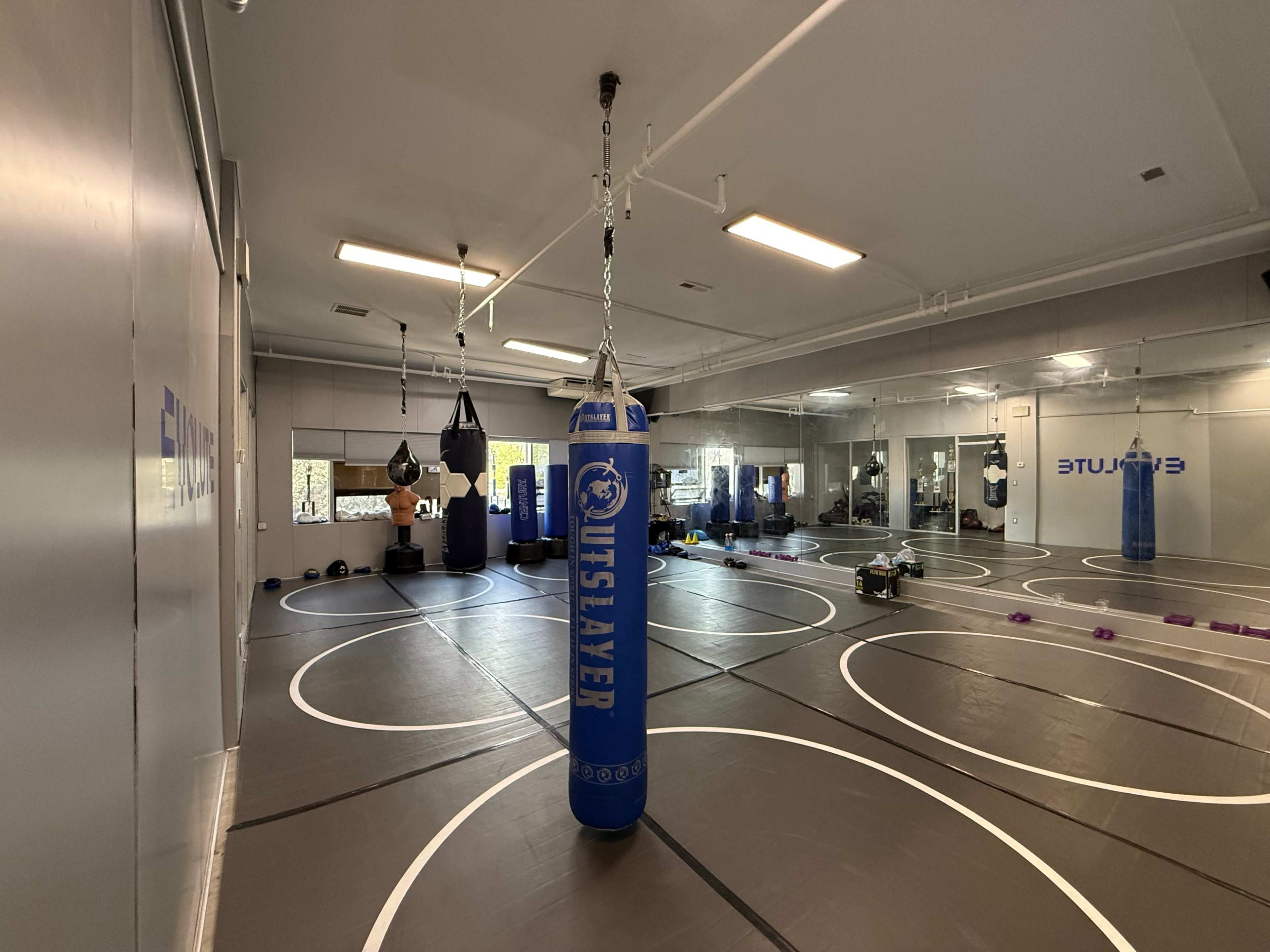 The image shows an indoor training facility with a blue punching bag in the center, surrounded by exercise mats and various workout equipment.