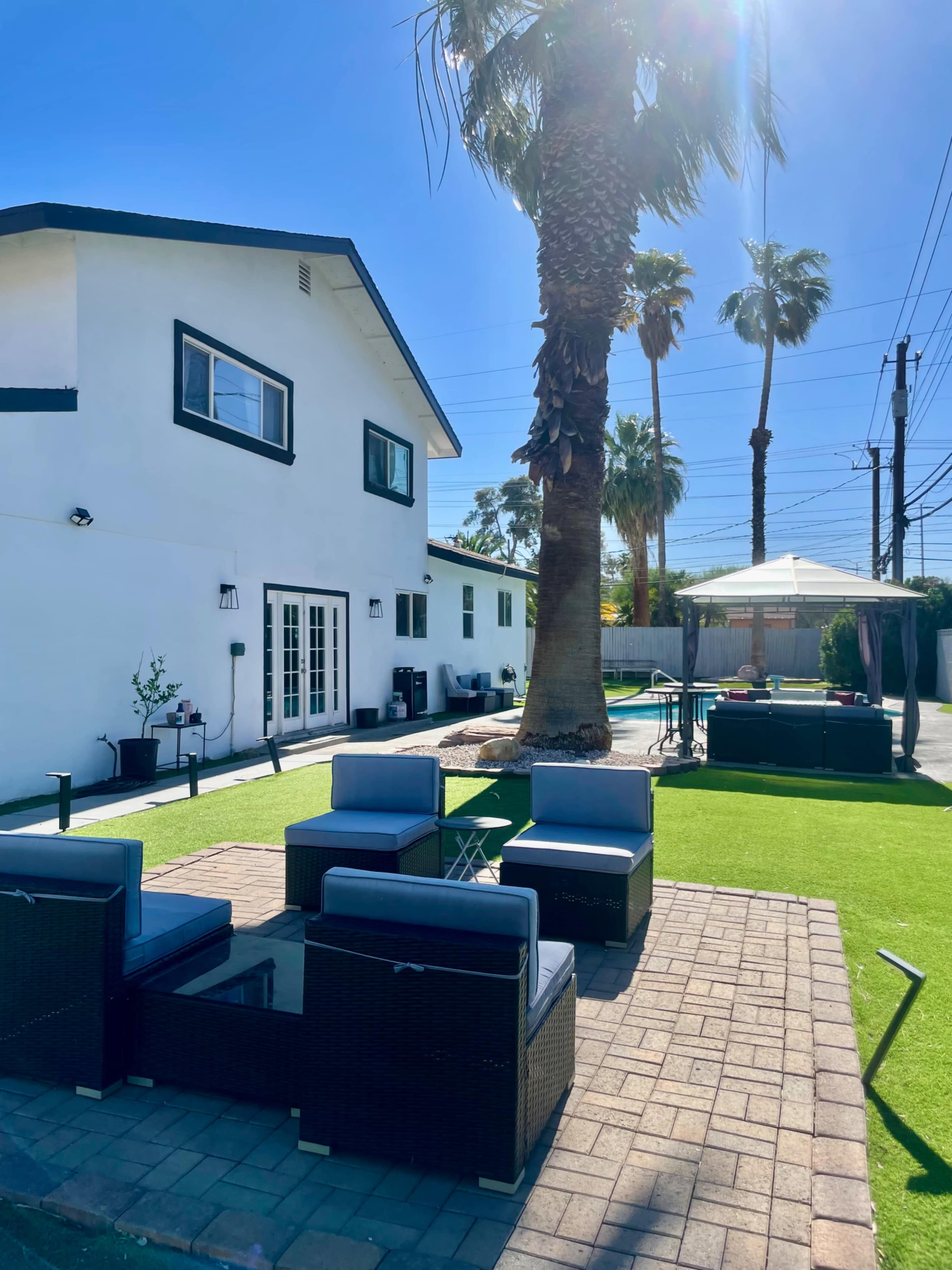 The image shows a backyard patio with outdoor seating, a palm tree, and a gazebo beside a house.