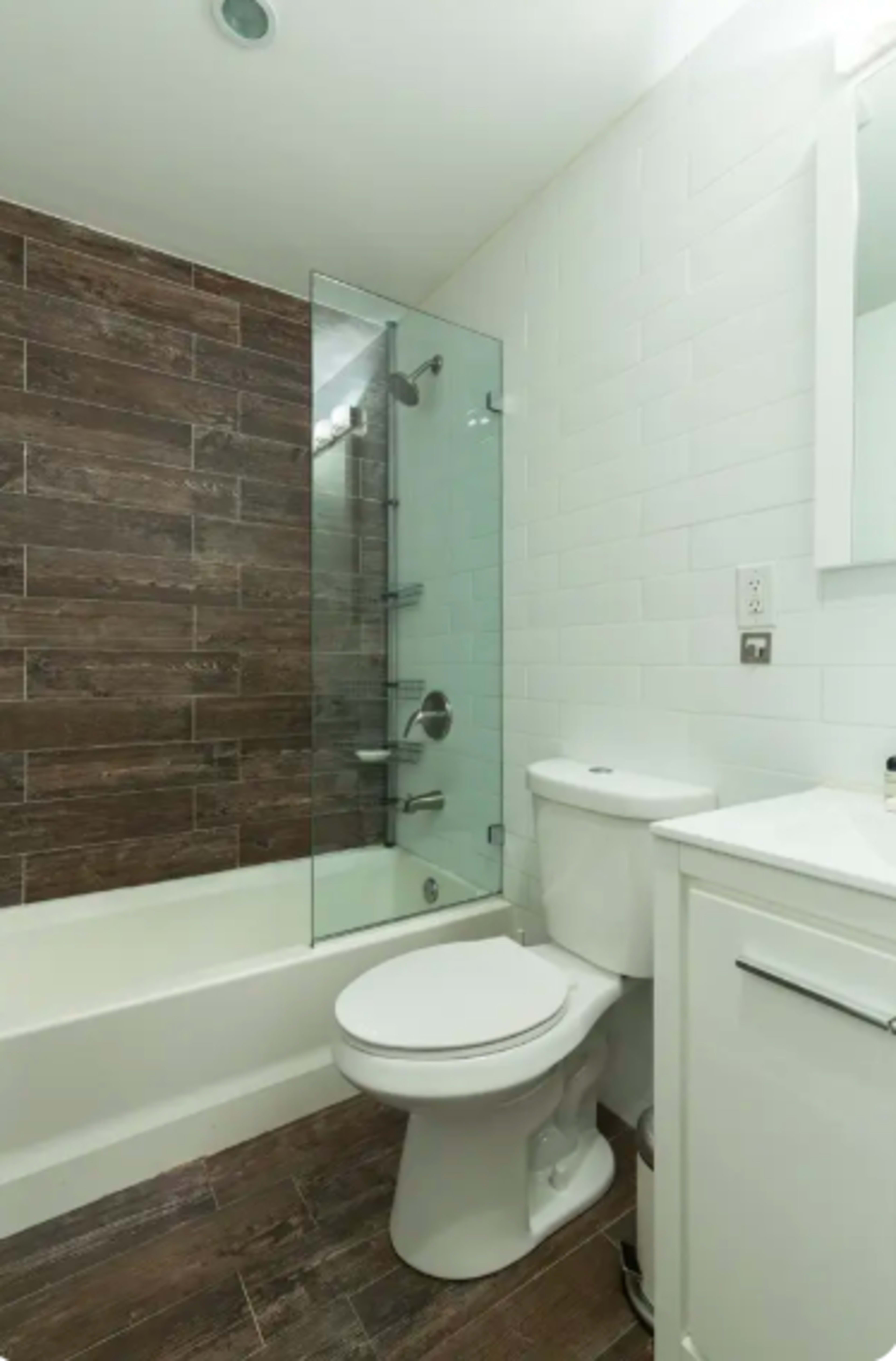 A modern bathroom featuring a shower with a glass panel, a bathtub, a toilet, and a white vanity with a mirror.