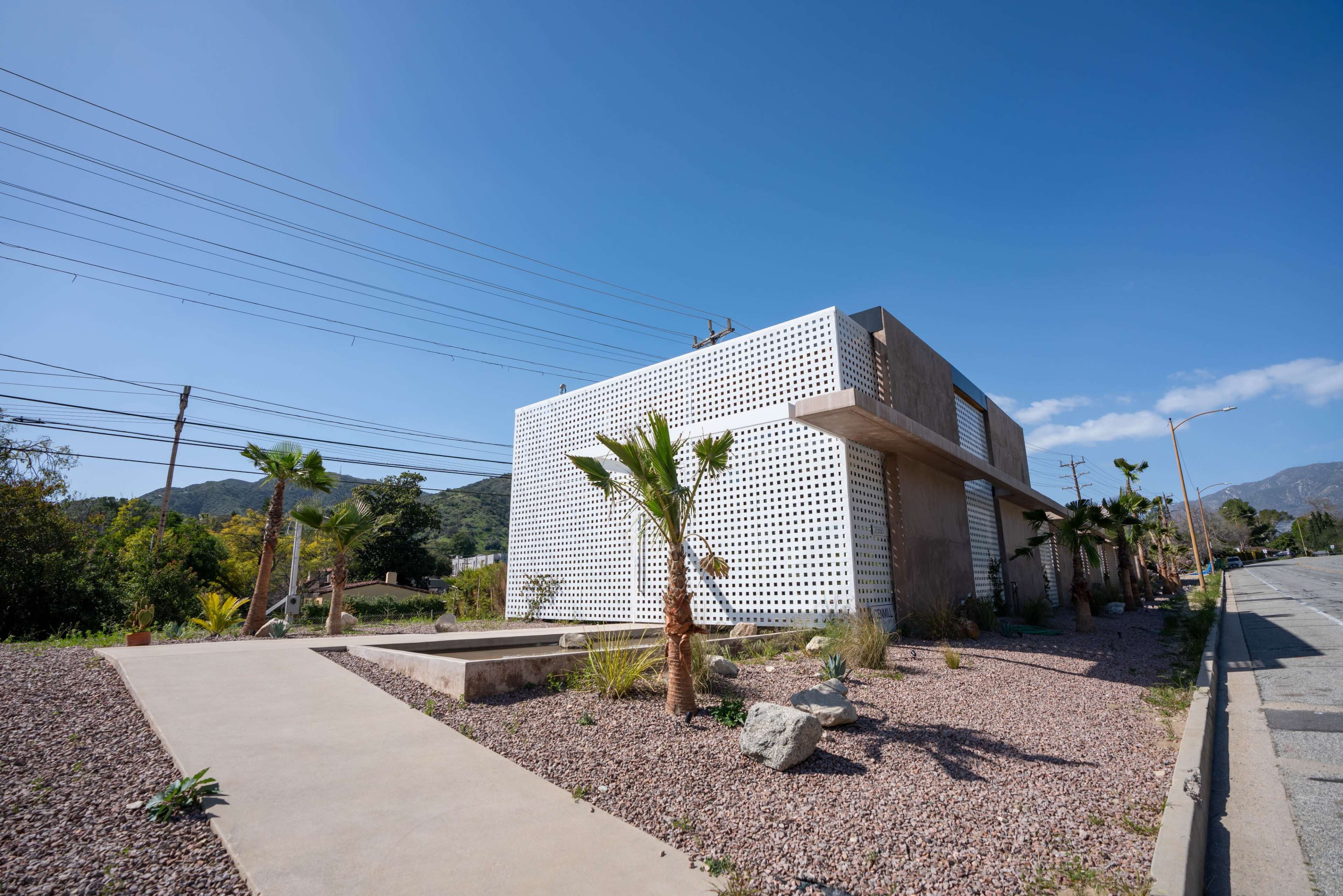 A modern building with a perforated facade is surrounded by landscaped stones and palm trees along a roadside.