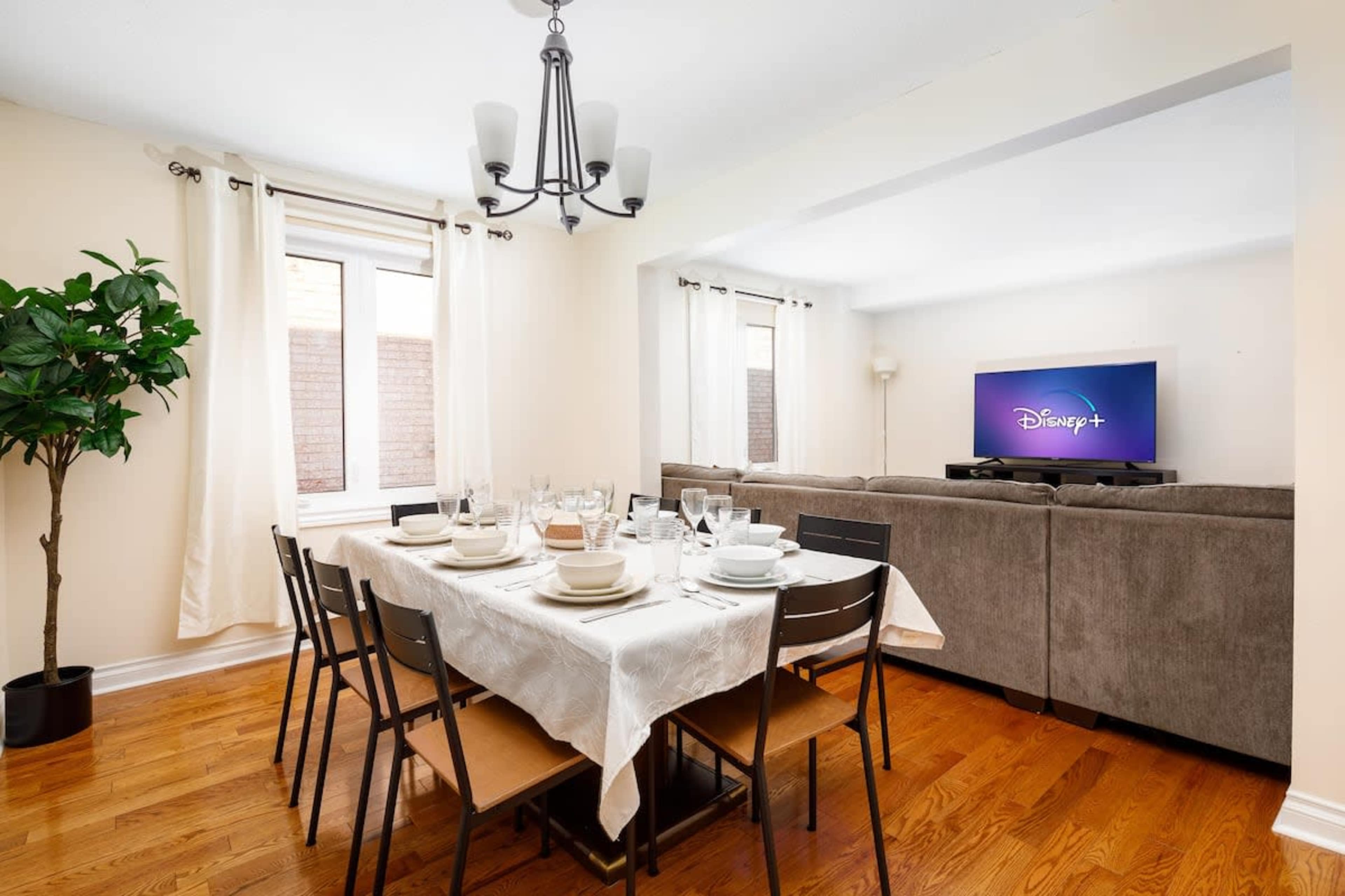A dining room features a table set for a meal, a television displaying the Disney+ logo, and a sectional couch in the background.
