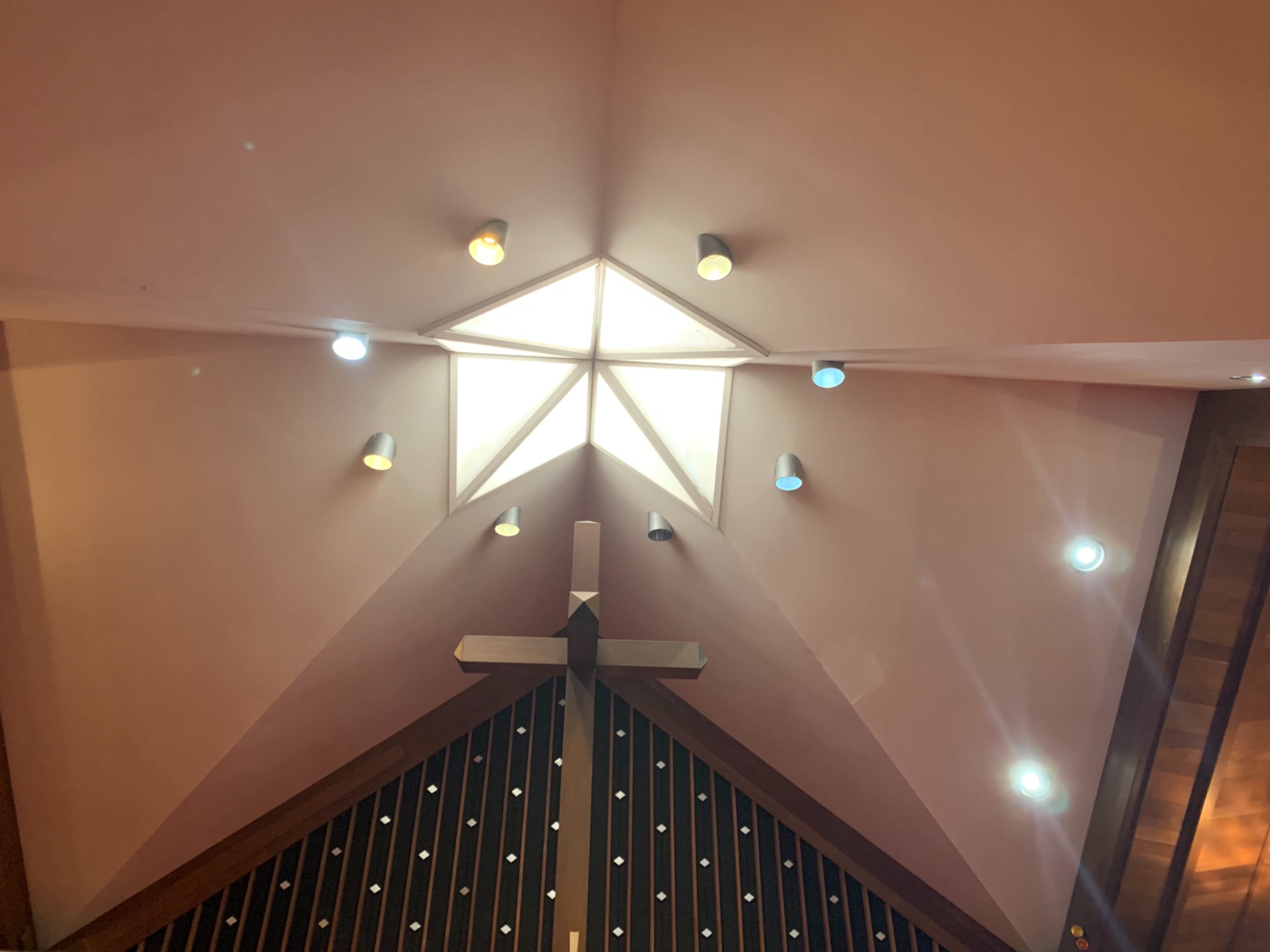 The image shows a church interior featuring a triangular ceiling with a central skylight and a wooden cross below it.