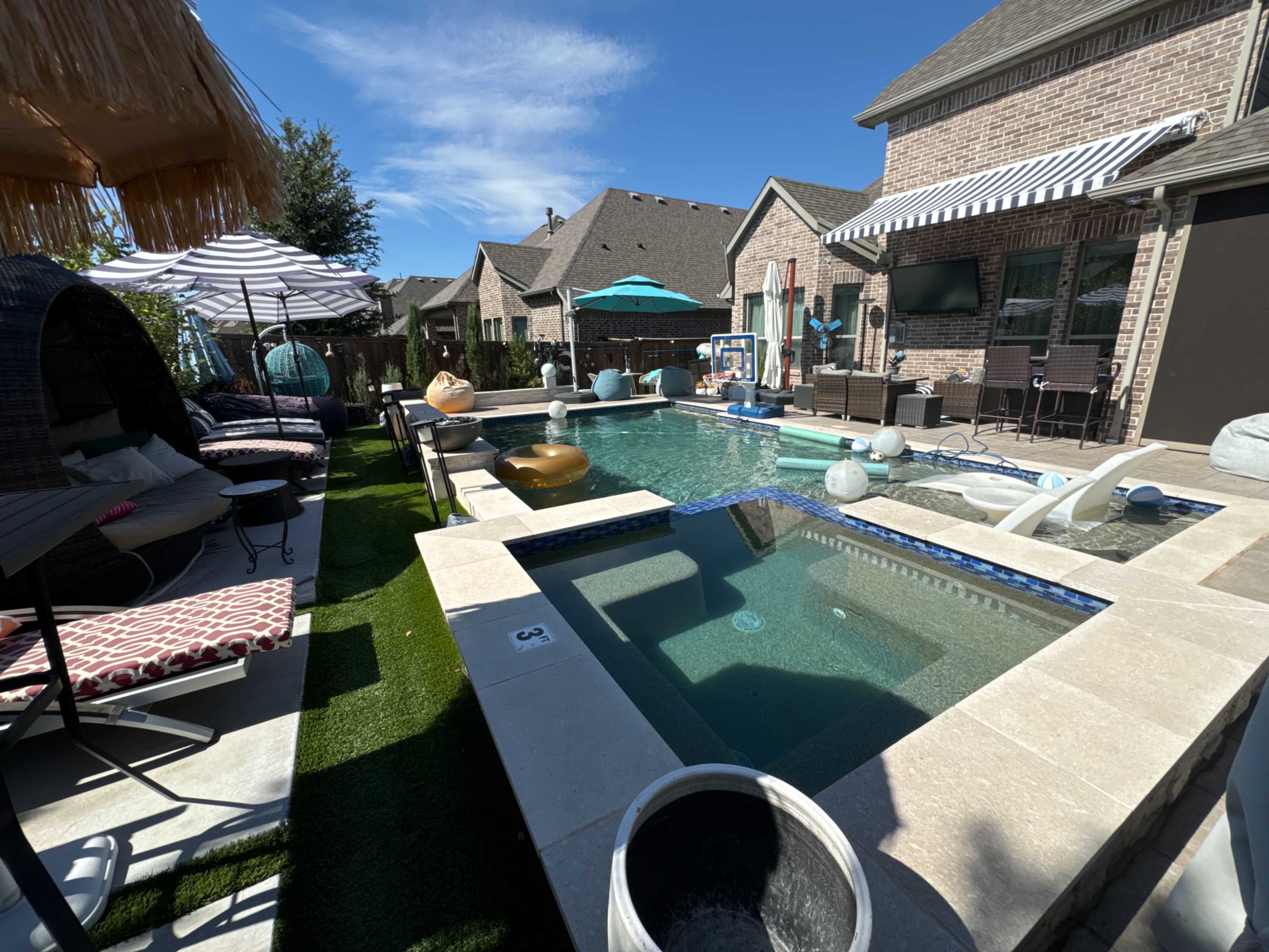 The image shows a residential backyard featuring a swimming pool with a hot tub, surrounded by lounge chairs and shaded seating areas.