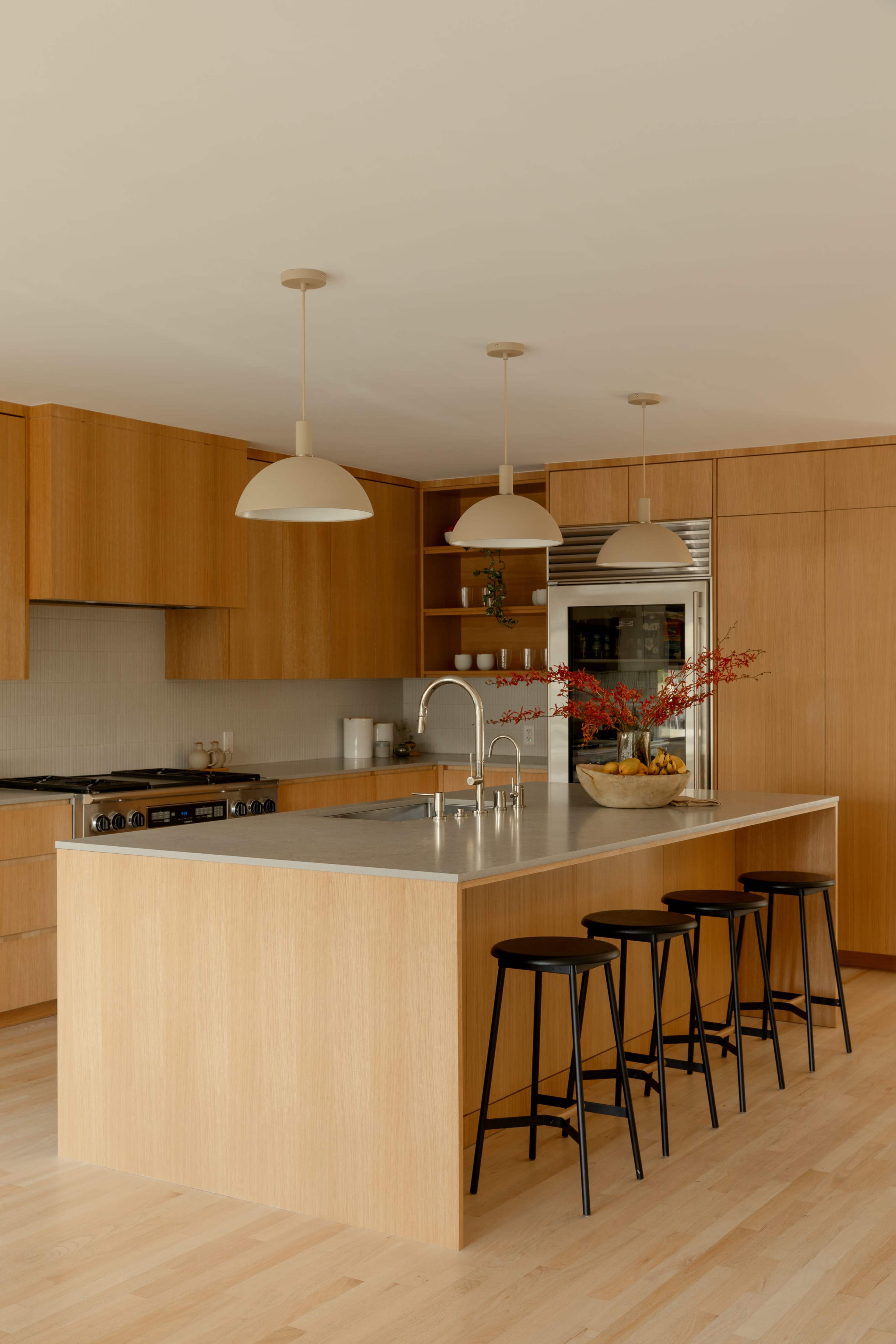 A modern kitchen features wooden cabinetry, a large island with a stainless steel countertop, and four black stools.