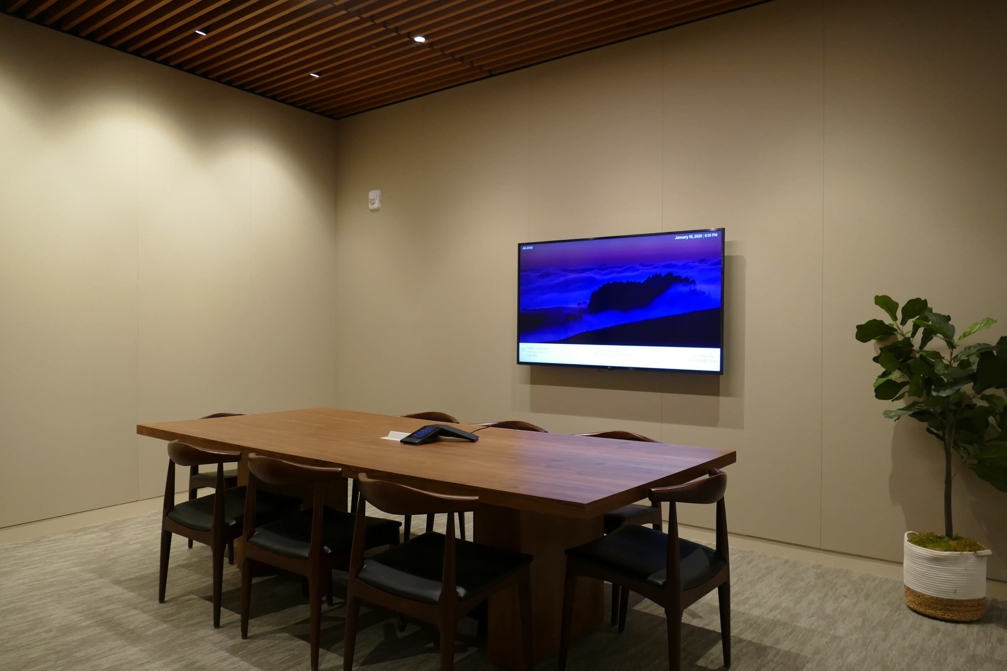 A conference room features a wooden table surrounded by chairs, with a wall-mounted television and a potted plant in the corner.