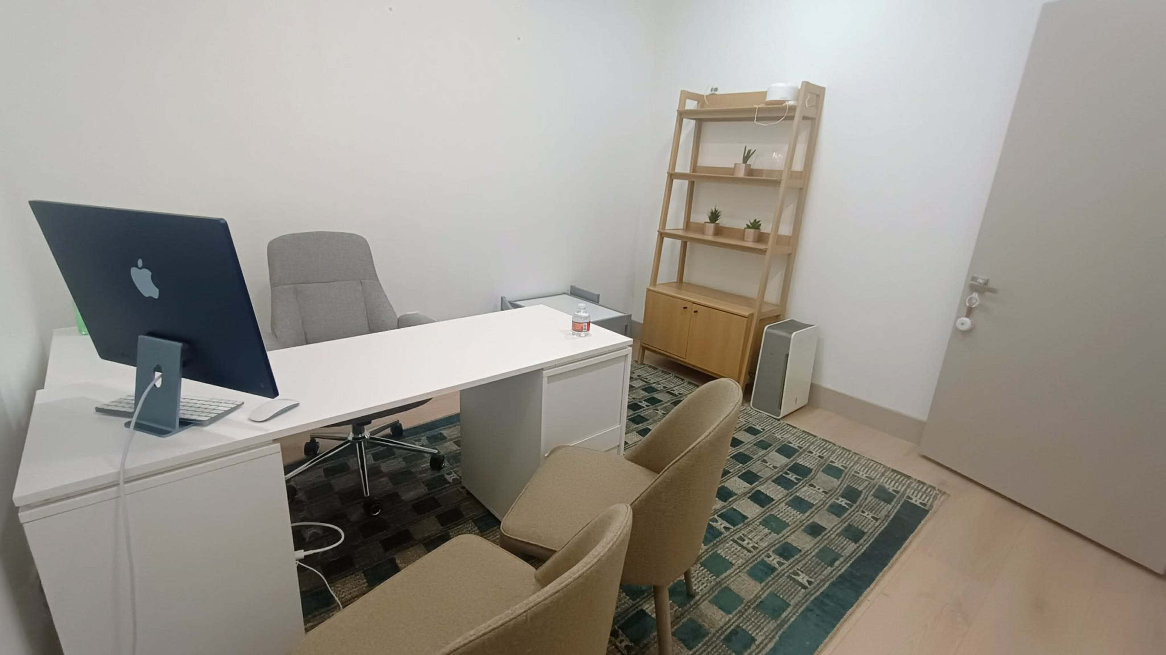 The image shows a minimalist office with a desk, an ergonomic chair, a computer, a bookshelf, and two chairs arranged on a patterned rug.