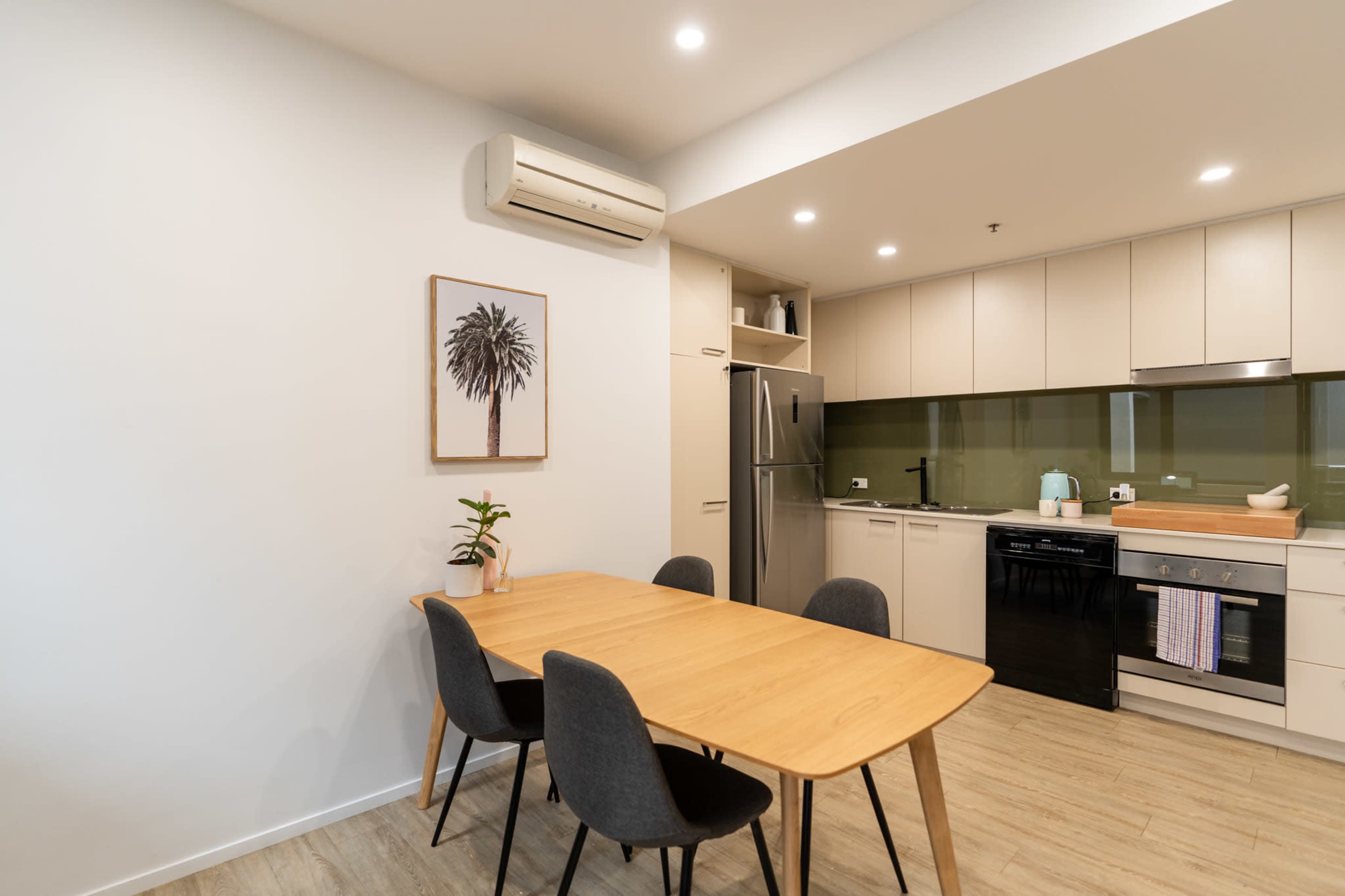 A modern kitchen and dining area features a wooden table with black chairs, an air conditioning unit, and sleek appliances against a green backsplash.