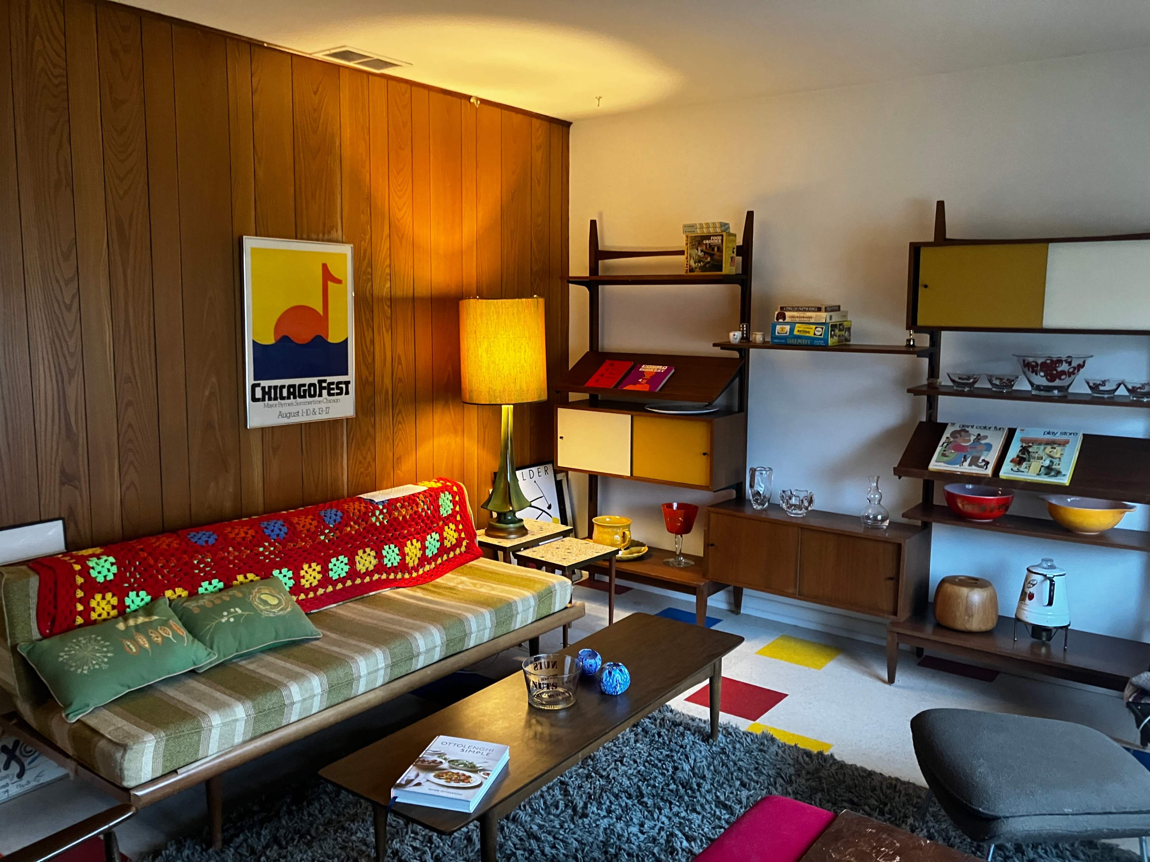 A mid-century modern living room features a sofa with a colorful crocheted blanket, a wooden bookshelf, and a retro lamp, surrounded by various decor items.