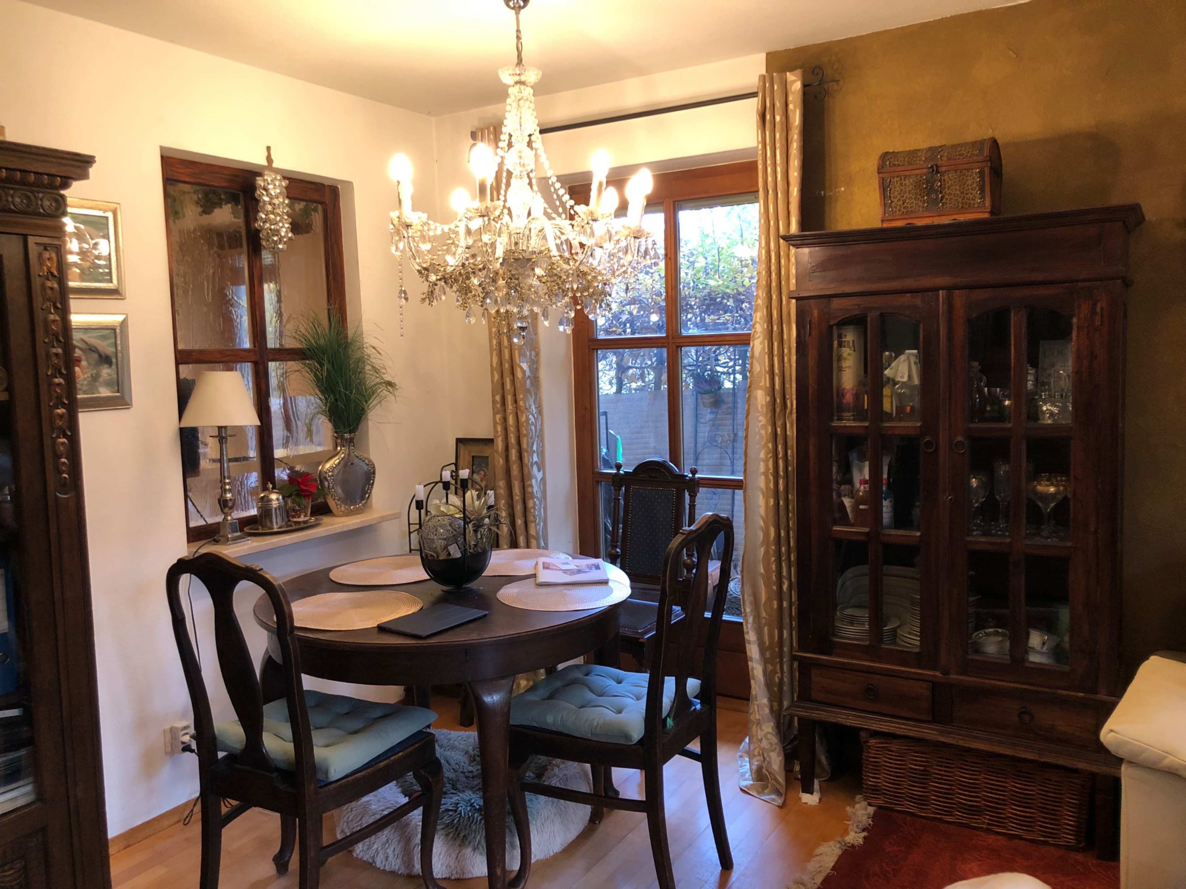 The room features a round dining table surrounded by wooden chairs, a chandelier overhead, and a glass-front cabinet displaying various items.
