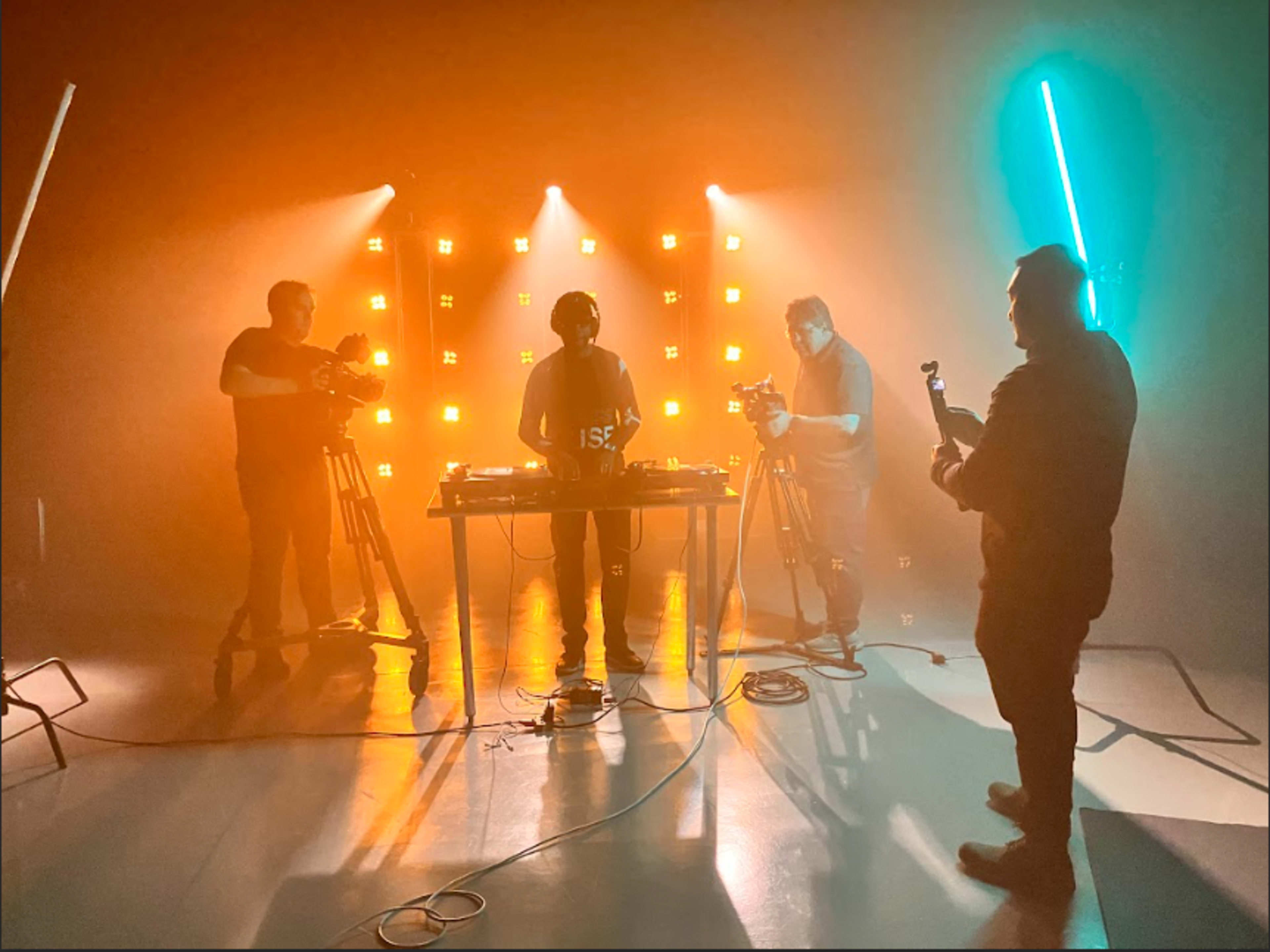 A DJ stands behind a table surrounded by three camera operators in a studio with bright, colored lights.