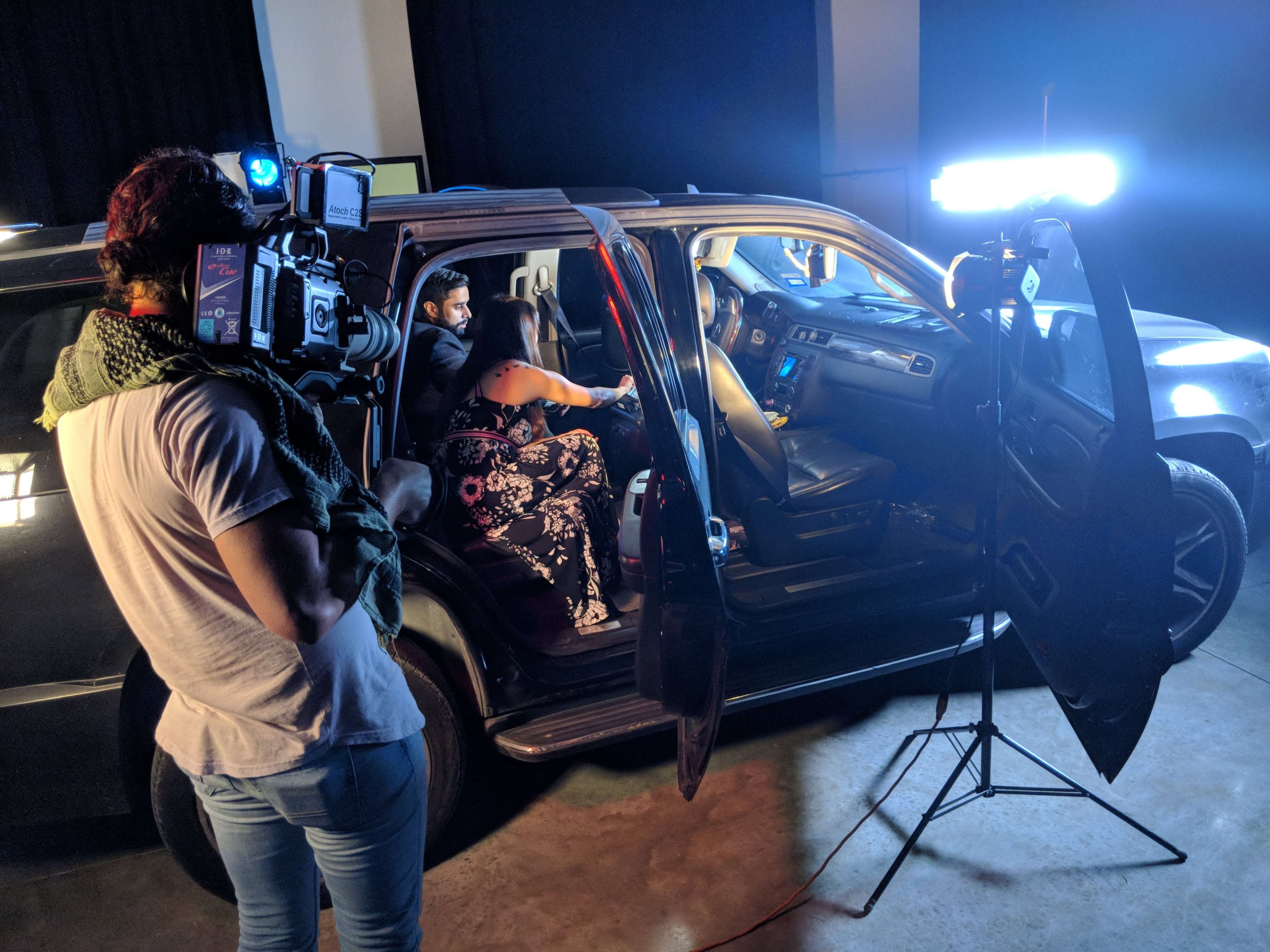 A film crew sets up to shoot a scene inside an SUV with actors seated in the front, illuminated by artificial lights.