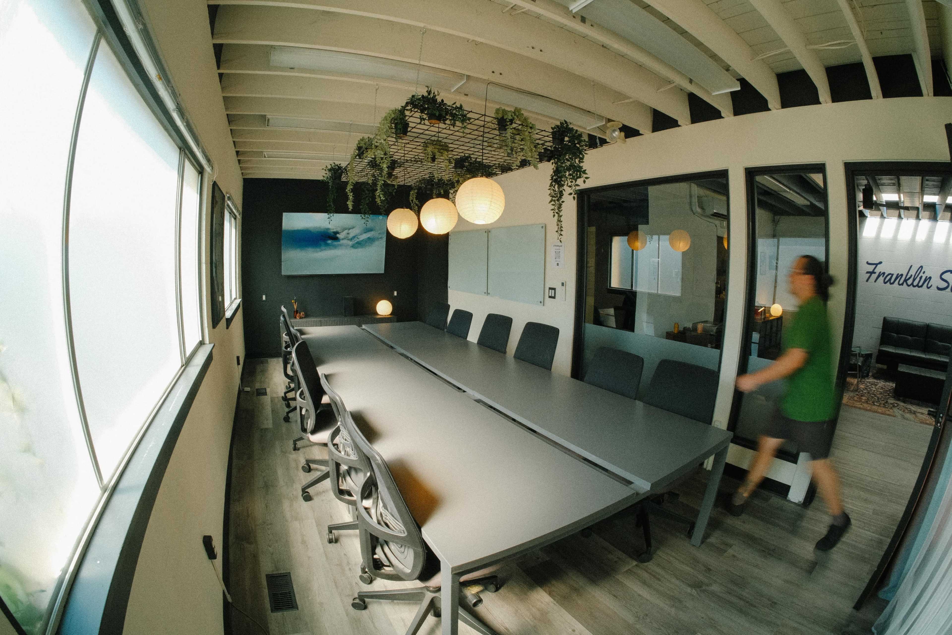 A spacious conference room features a long table surrounded by chairs and illuminated by hanging lights and greenery.