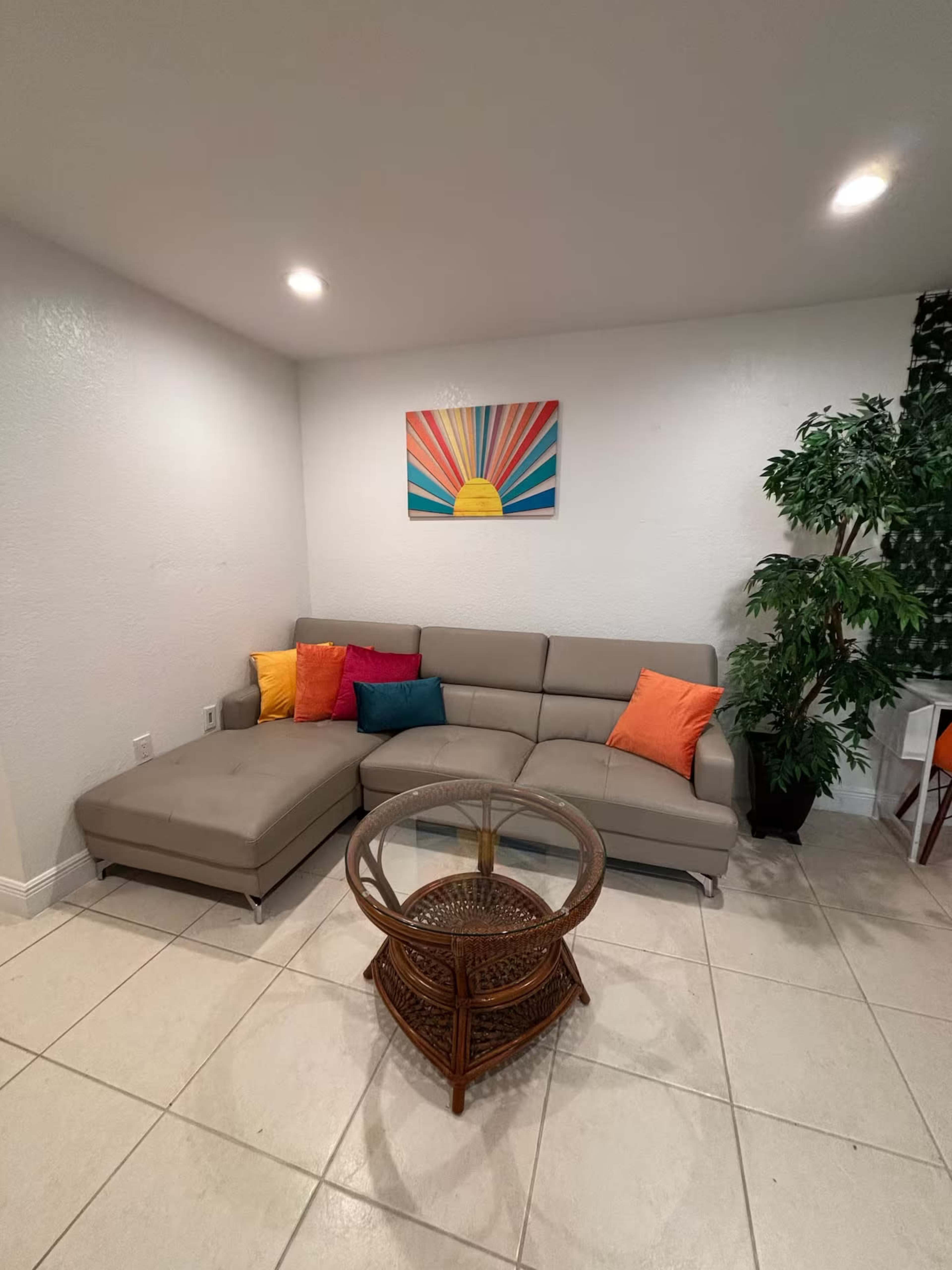 The image shows a cozy living area with a sectional sofa, colorful throw pillows, a round glass coffee table, and a potted plant against a light-colored wall, featuring a sunburst artwork above the sofa.