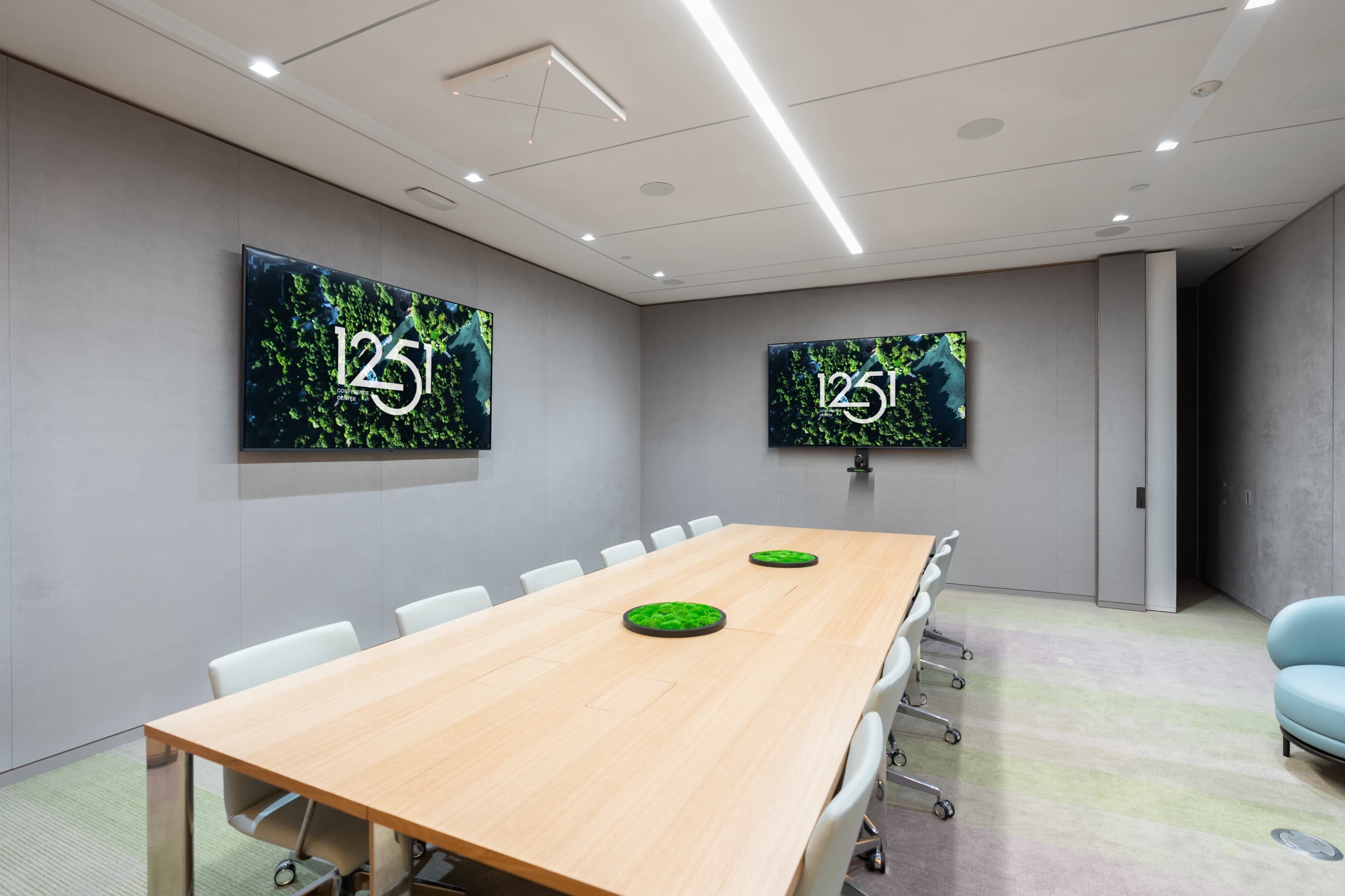 A modern conference room features a long wooden table, white chairs, and two large screens displaying a green-themed graphic.