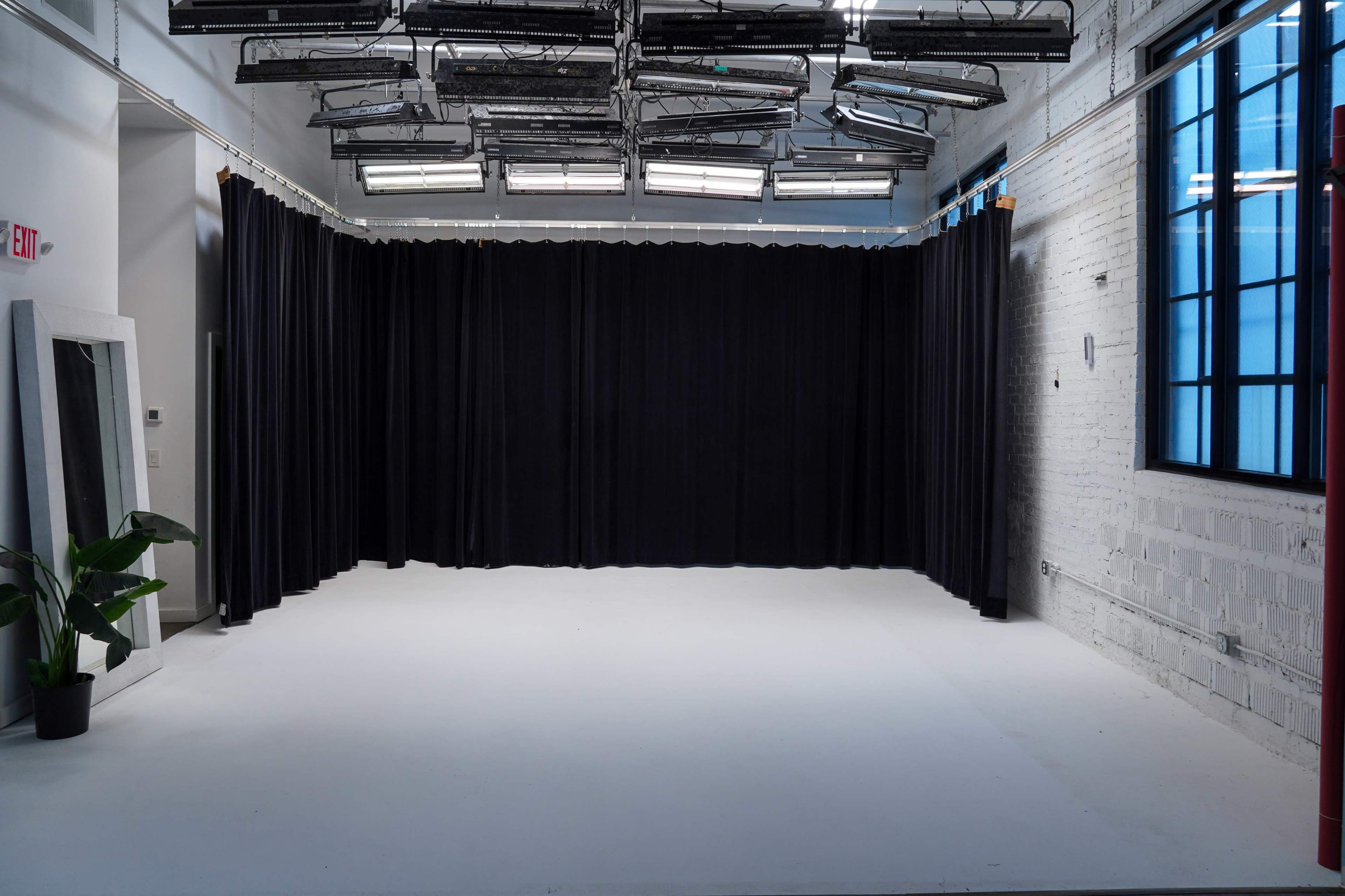 The image shows a spacious room with a white floor, black curtains on the left side, and large windows on the right.