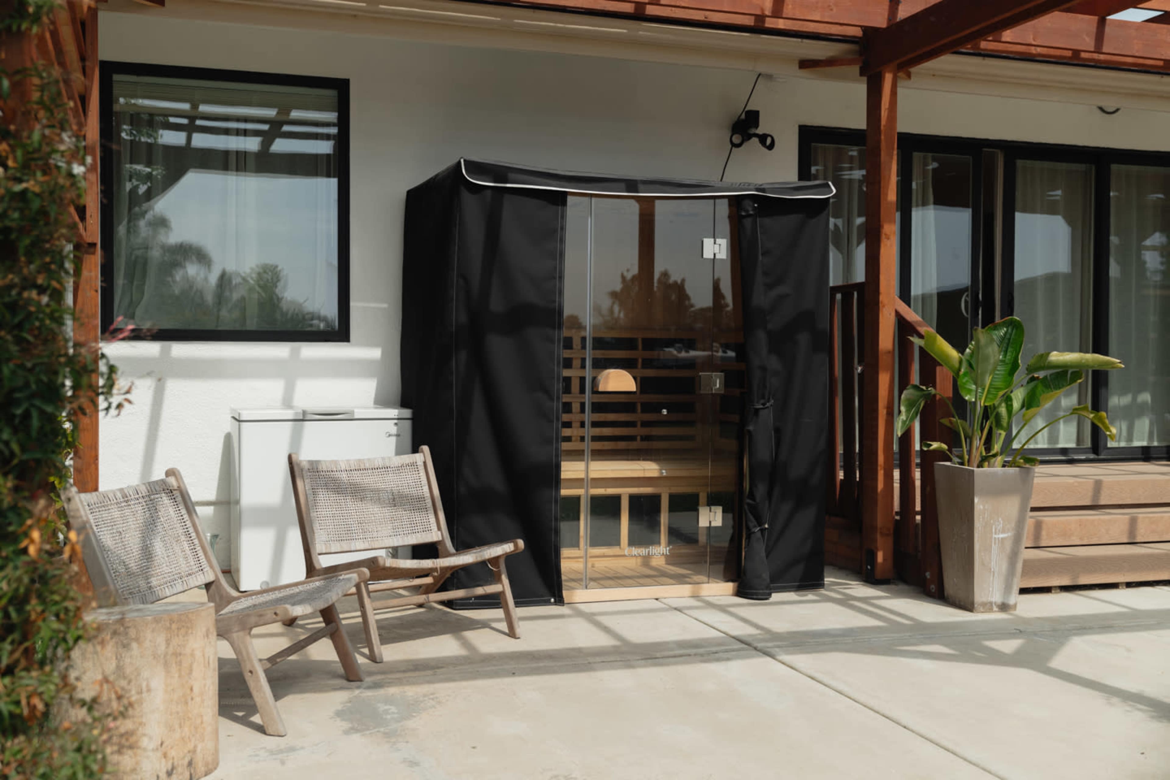 A black sauna is positioned next to two wooden chairs on a concrete patio beside a house.