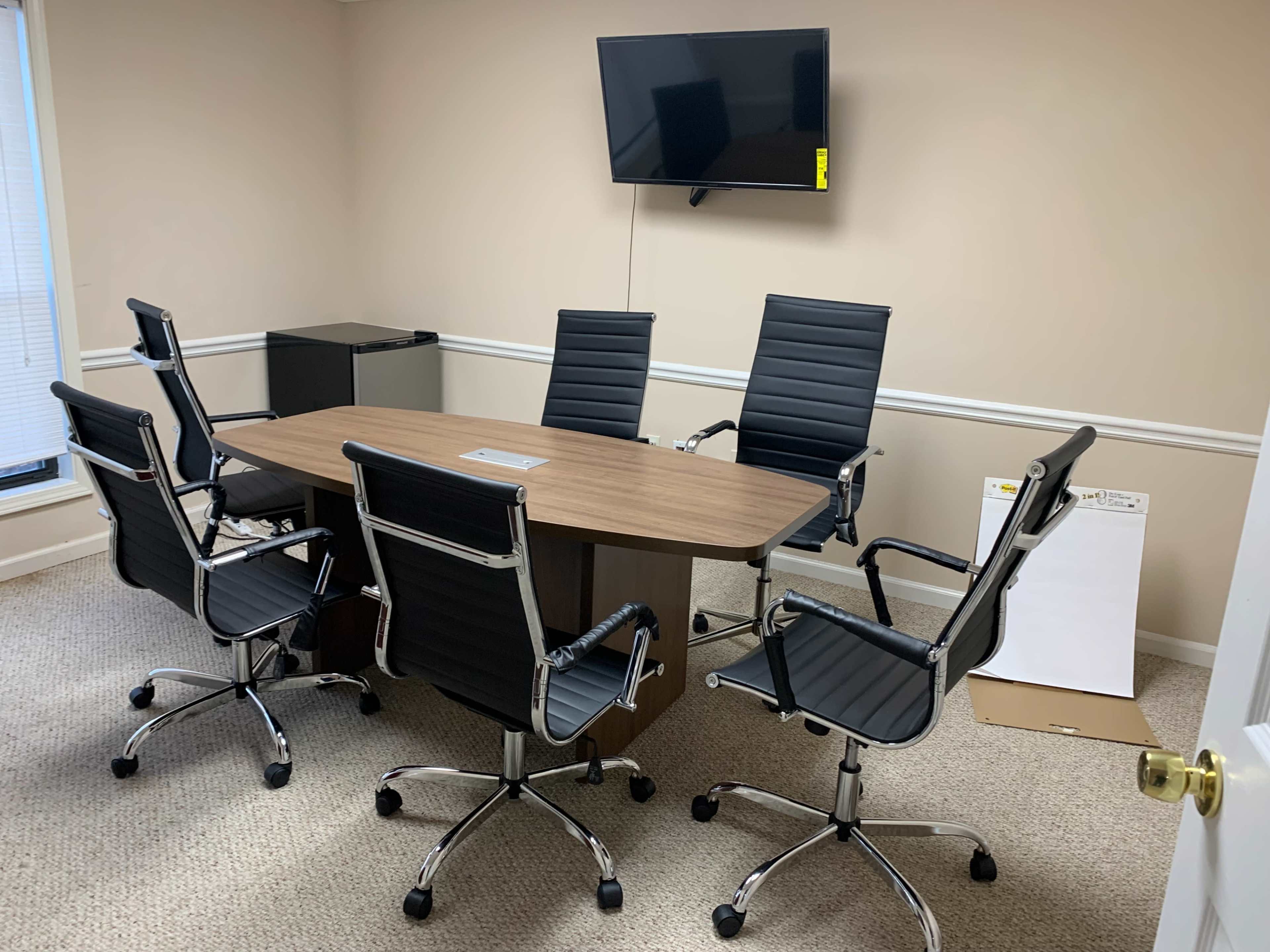 The image shows a clean, small conference room with a wooden table surrounded by six black office chairs and a wall-mounted television.