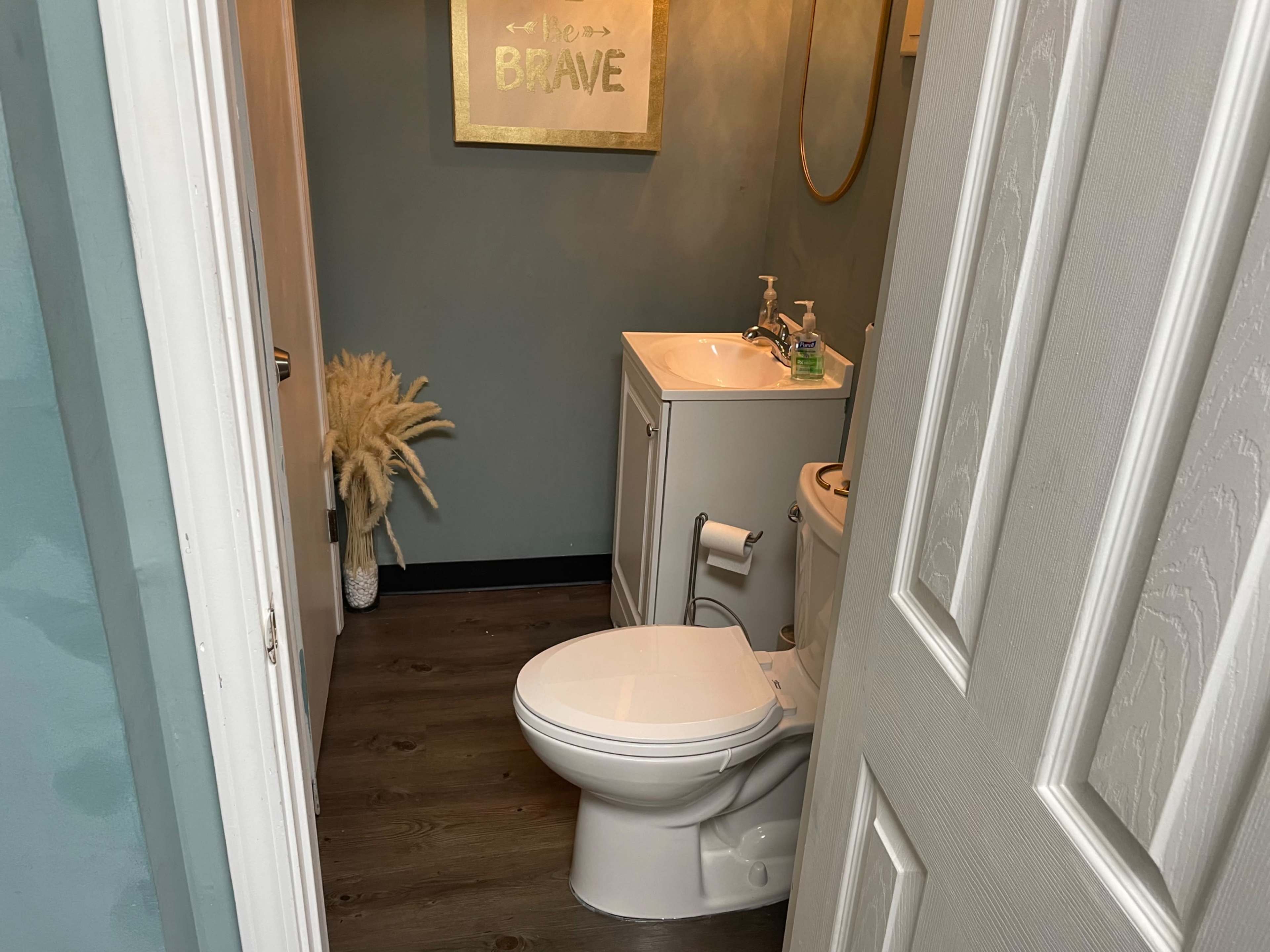 The image shows a small bathroom with a toilet, sink, and decorative wall art, accessible through a door.