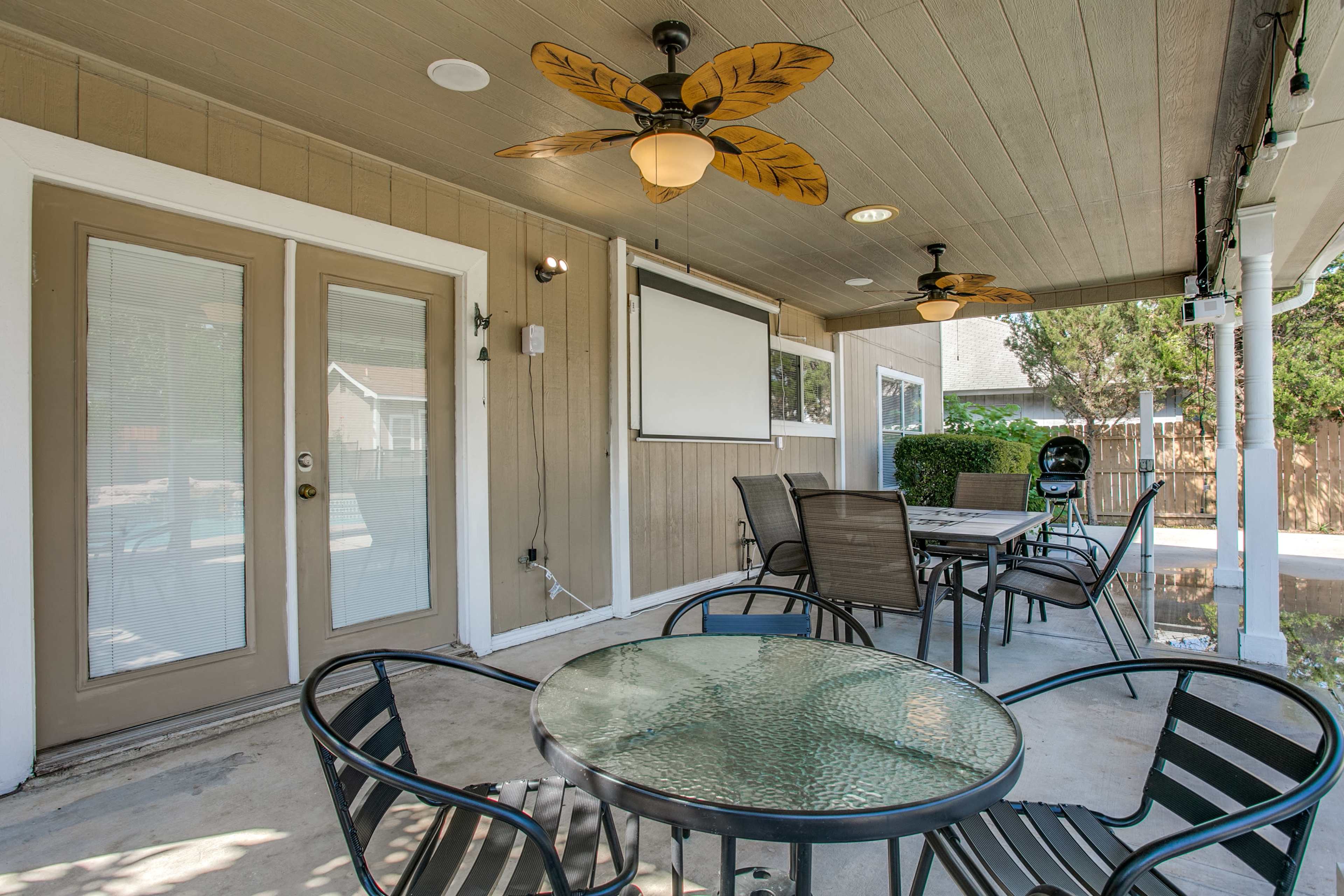A covered outdoor patio with a glass table and several chairs, featuring a ceiling fan and a wall-mounted screen.
