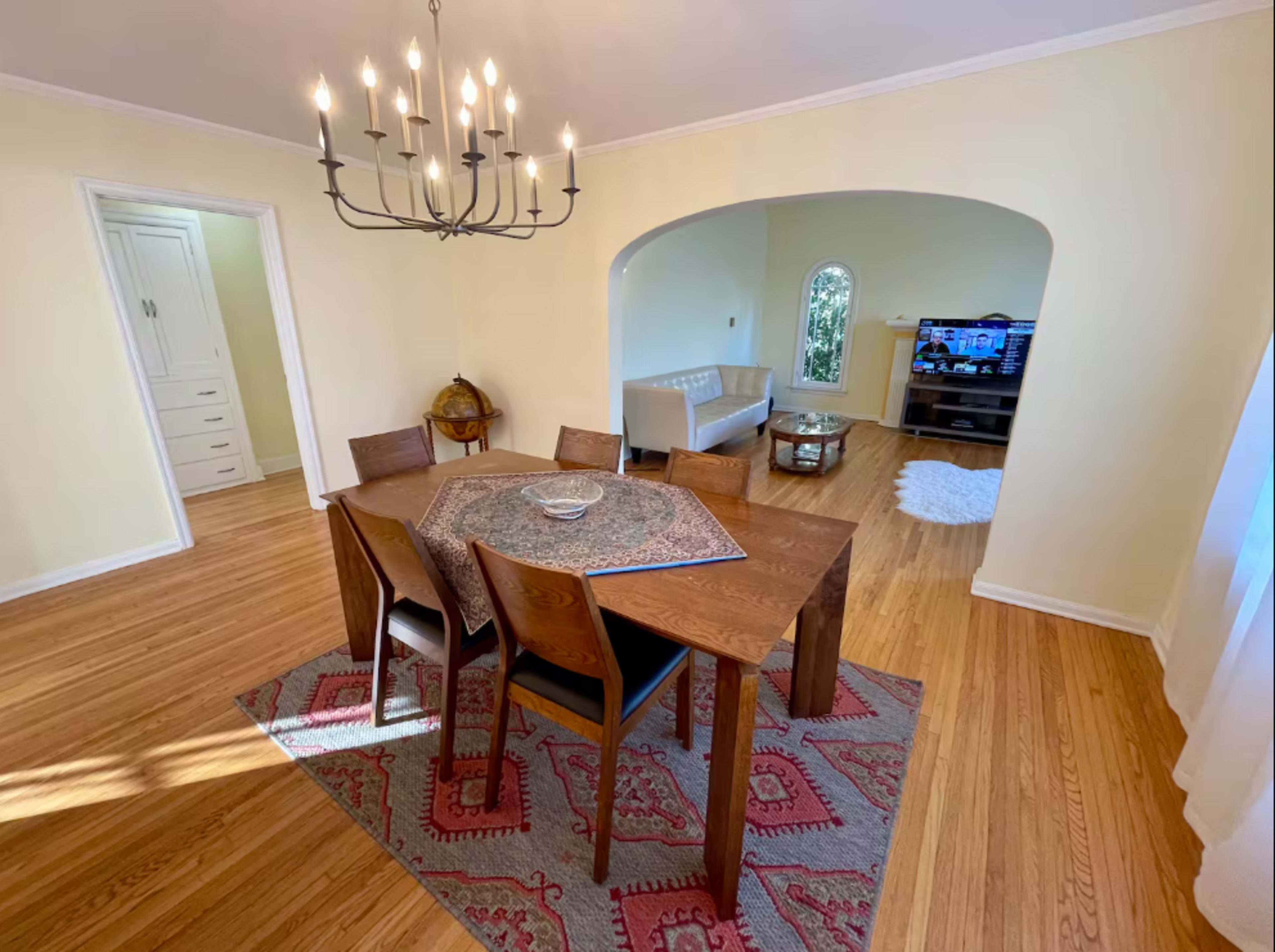 The image shows a dining area with a wooden table and chairs, a chandelier overhead, and an adjacent living space featuring a couch and a television.