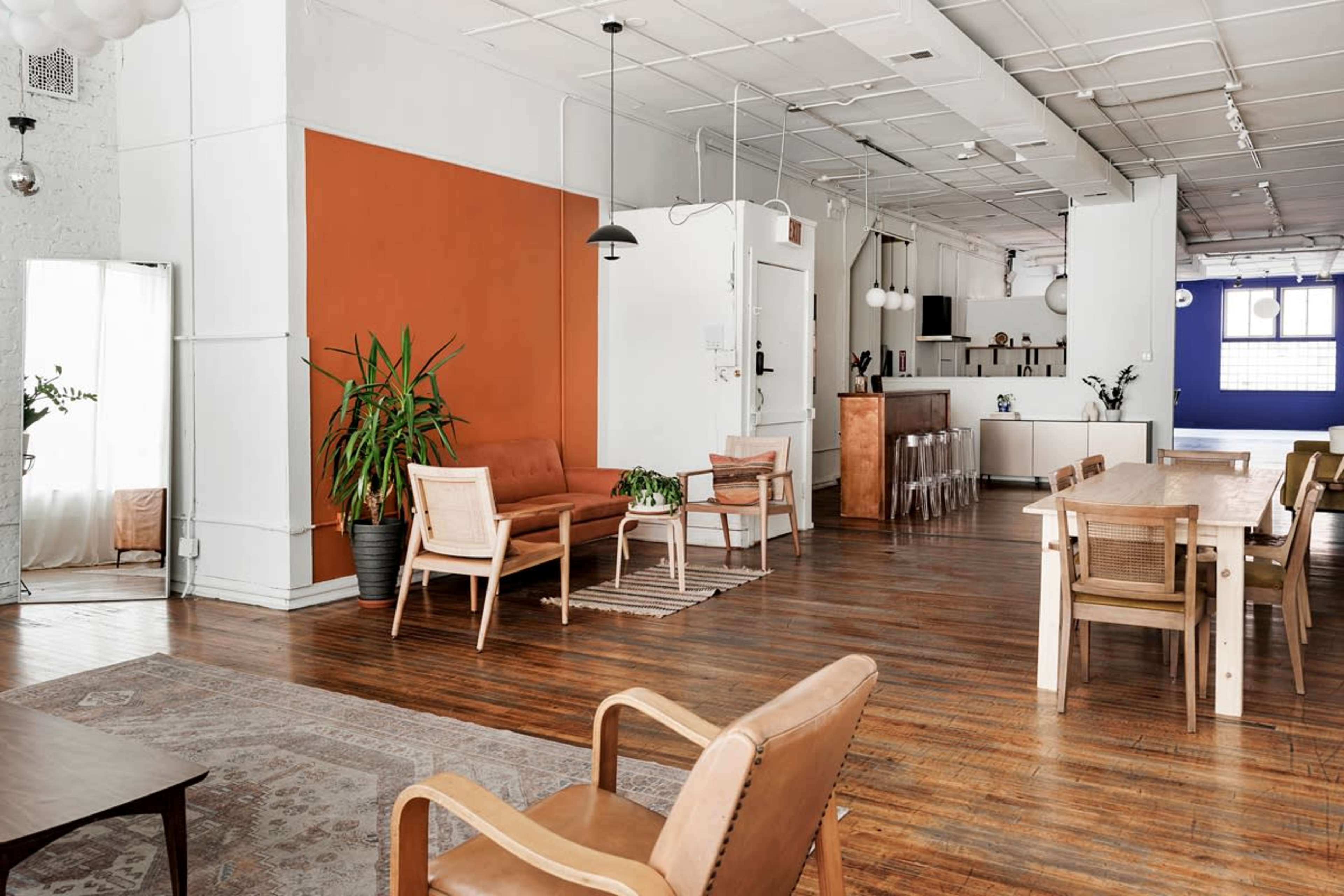 The image depicts a modern, open interior space featuring a seating area with brown furniture, plants, and a large dining table.