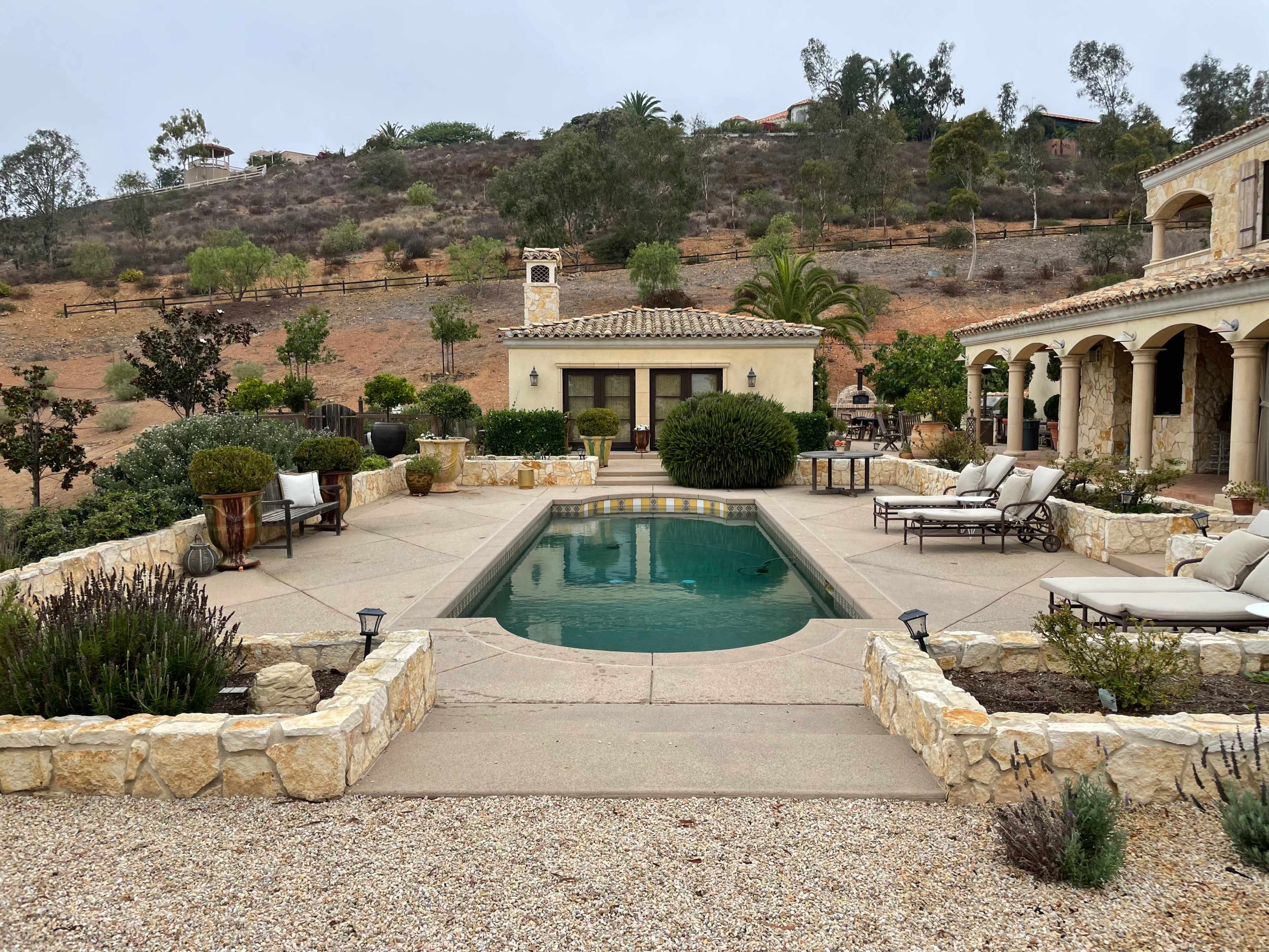 A landscaped outdoor area featuring a swimming pool surrounded by stone patios, lounge chairs, and various plants, set against a hillside.