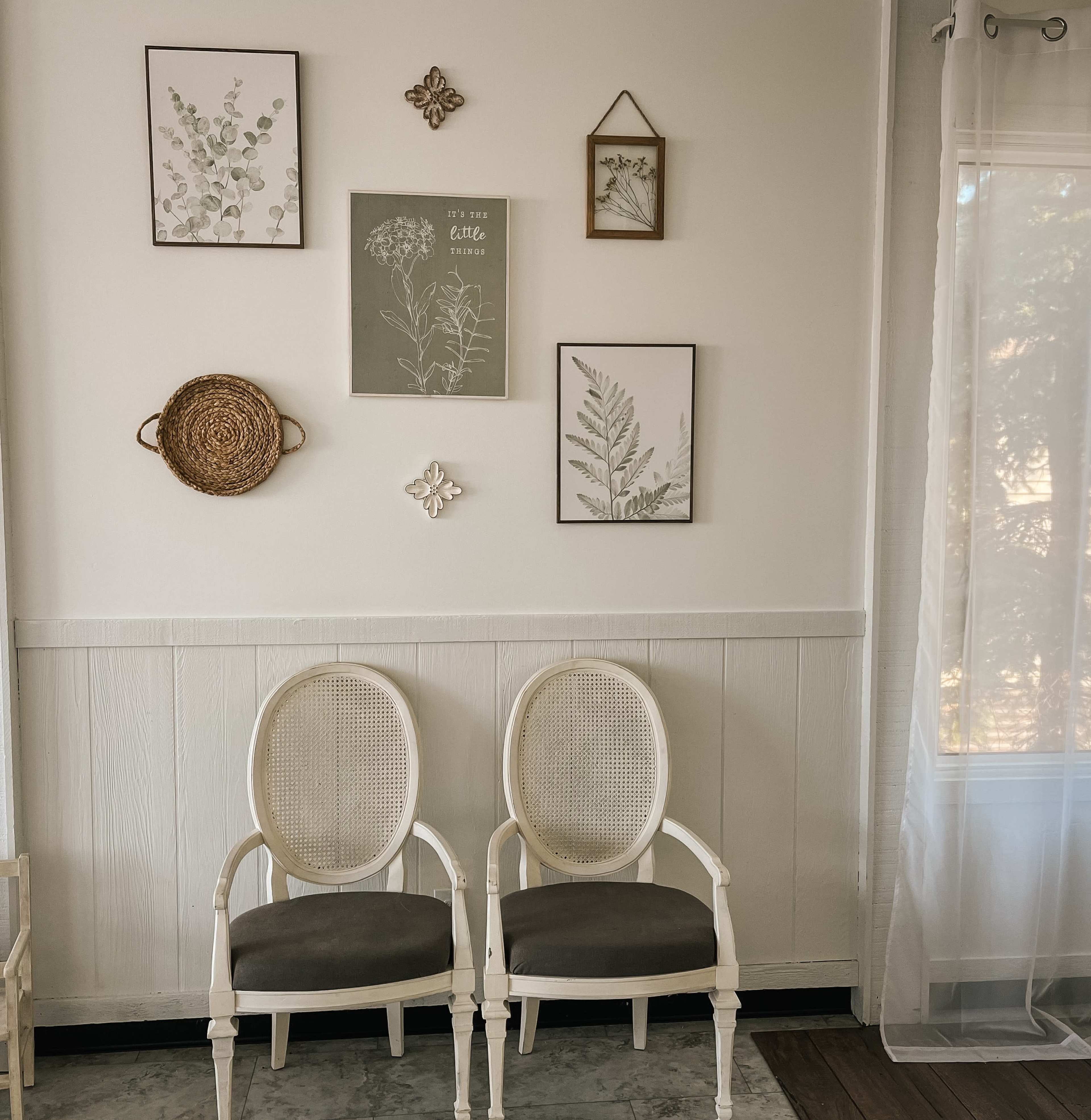 Two white chairs with gray cushions sit in front of a wall adorned with framed botanical prints and decorative items.