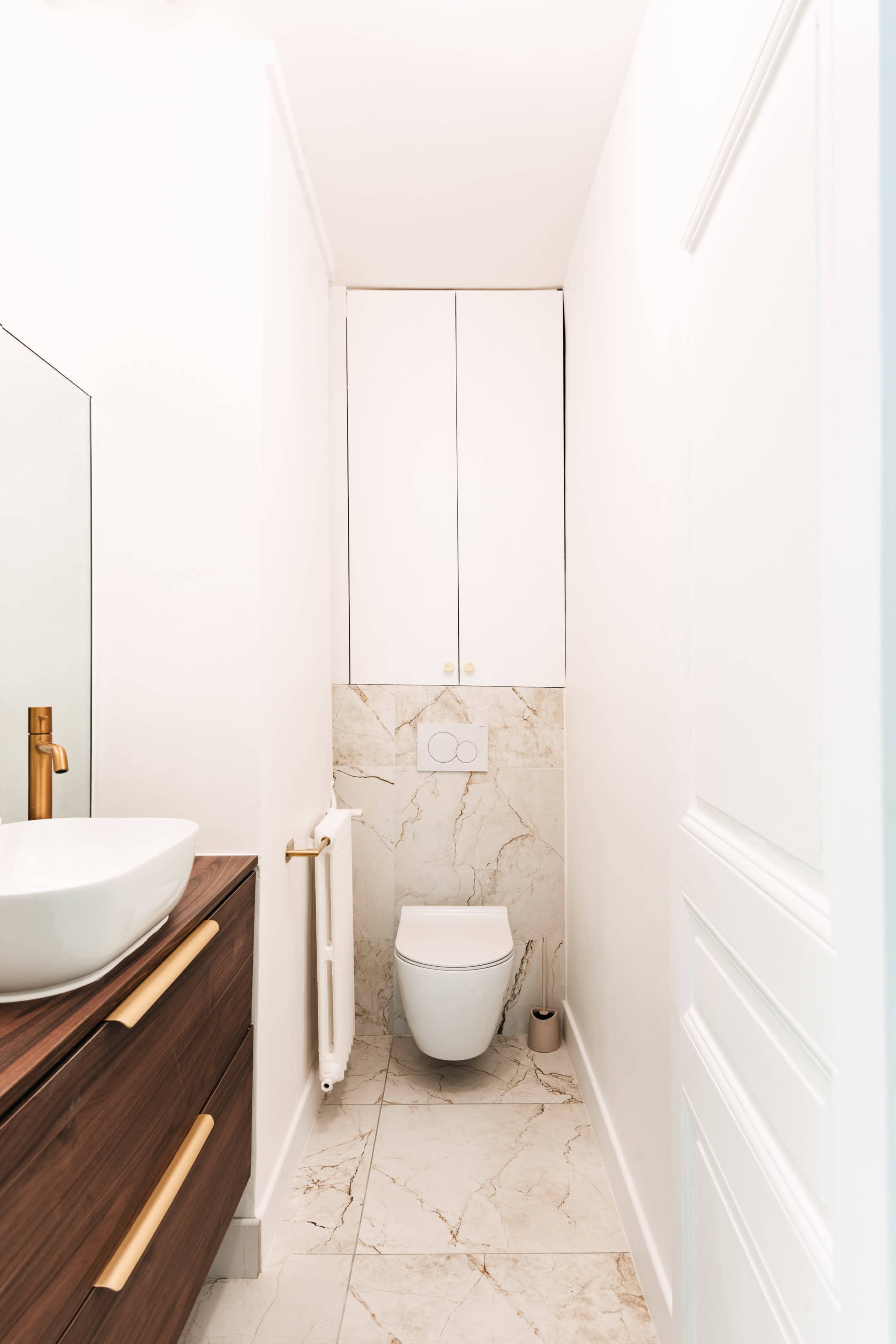 The image shows a narrow bathroom featuring a wall-mounted toilet, a sink with a wooden vanity, and white walls with marble accents.