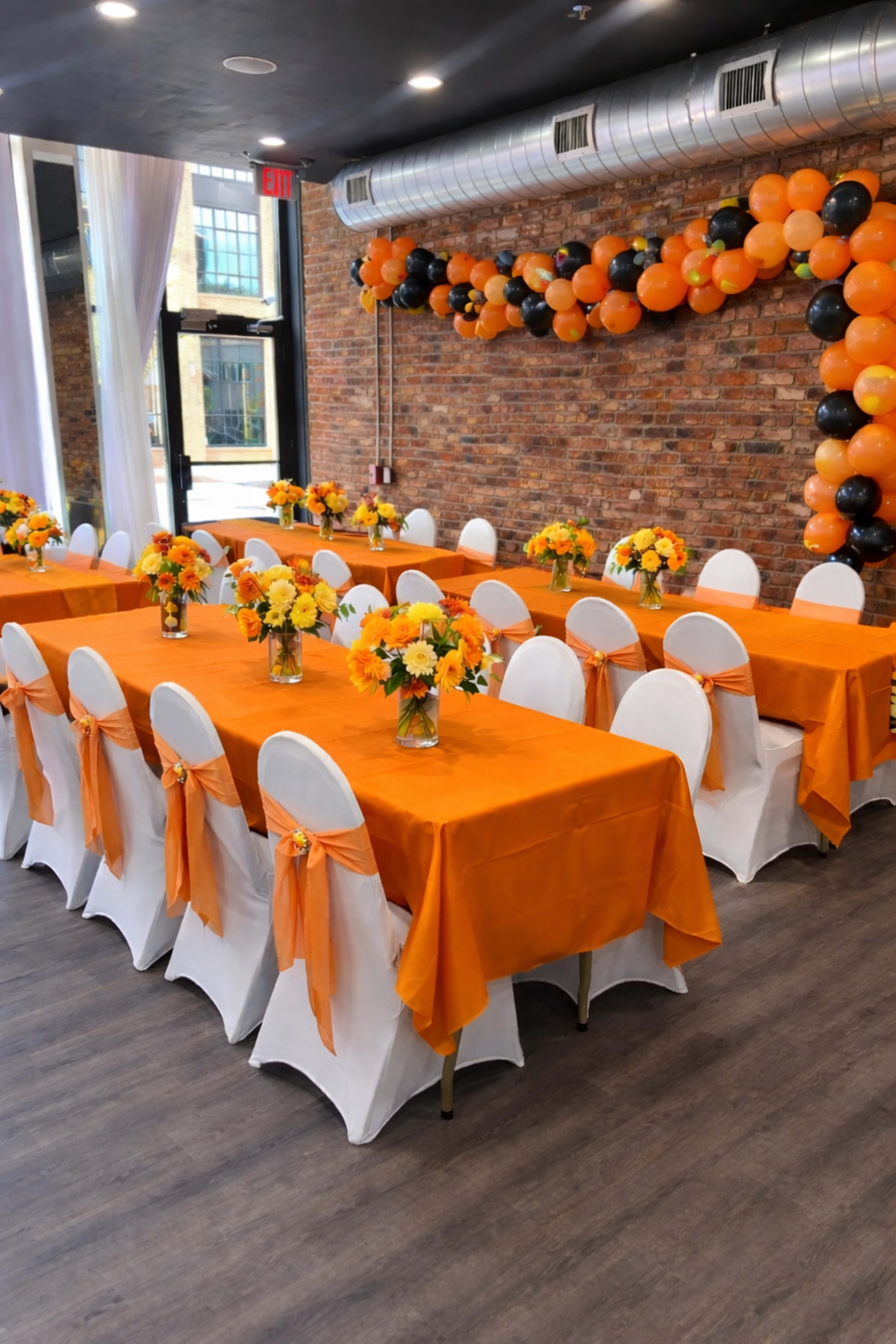 A banquet room features orange tablecloths and white chairs adorned with orange sashes, complemented by floral centerpieces and a balloon arch in shades of orange and black.