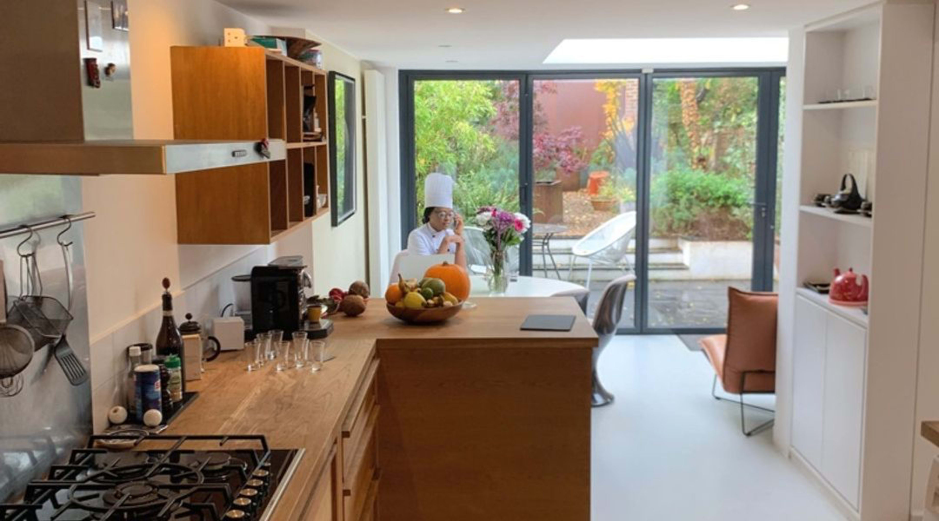 A modern kitchen opens up to a bright dining area with a view of a garden, featuring a person seated at a table surrounded by plants and natural light.