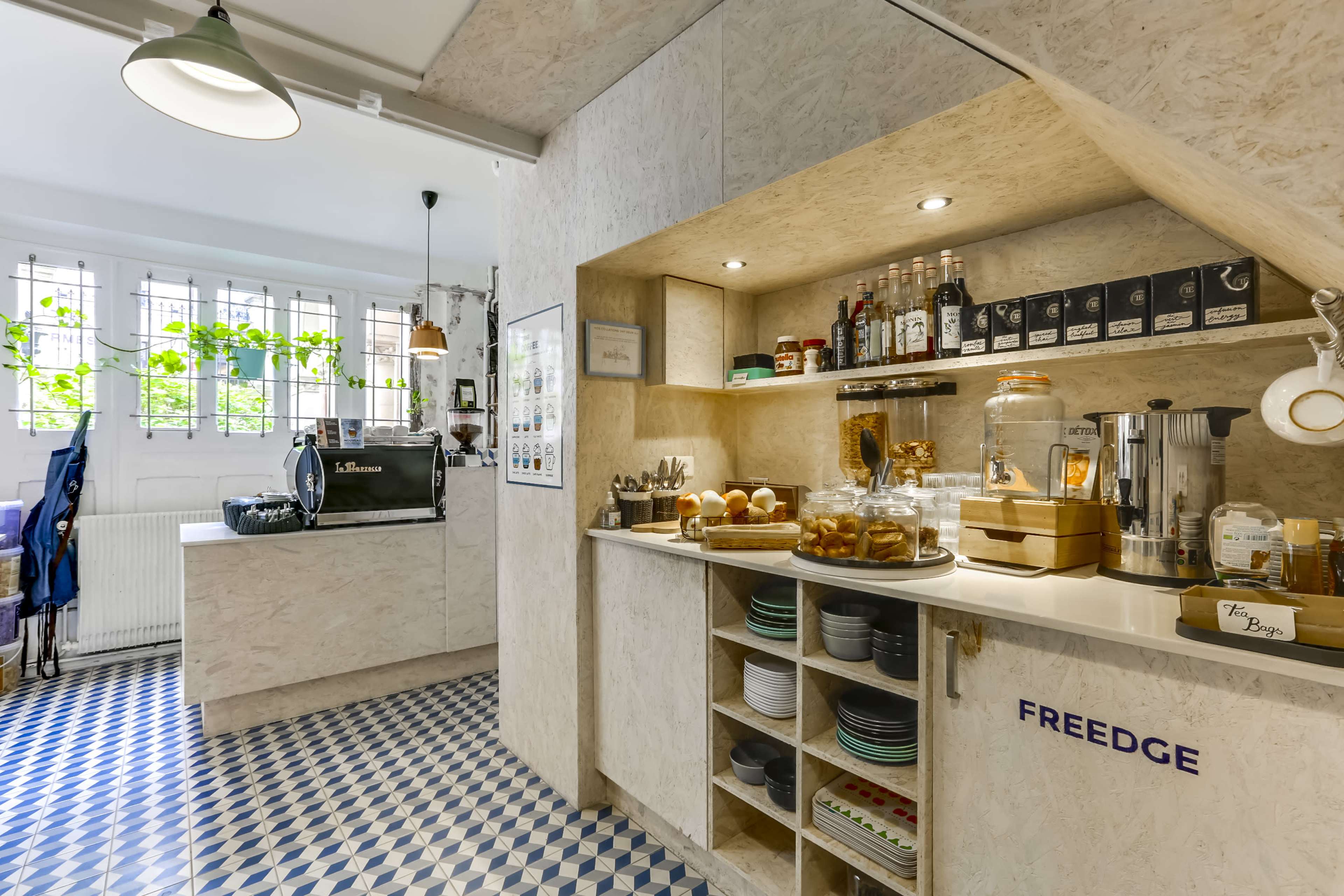 A modern café interior featuring a service counter with glass jars of food, shelves for drinks, and a coffee machine in an inviting space with patterned tile flooring.